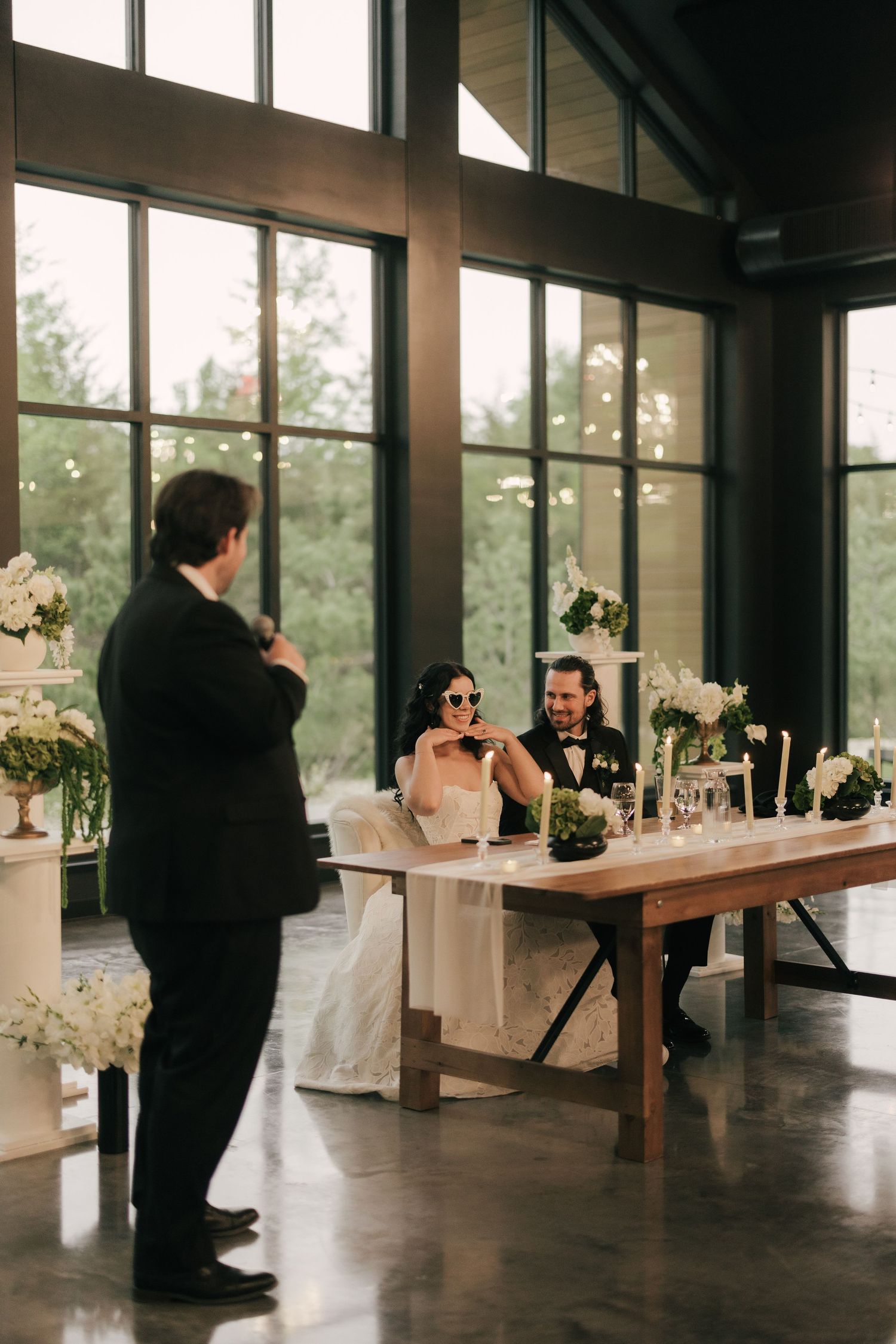 Bride and groom seated at sweetheart table during wedding reception toast inside elegant venue with large windows.