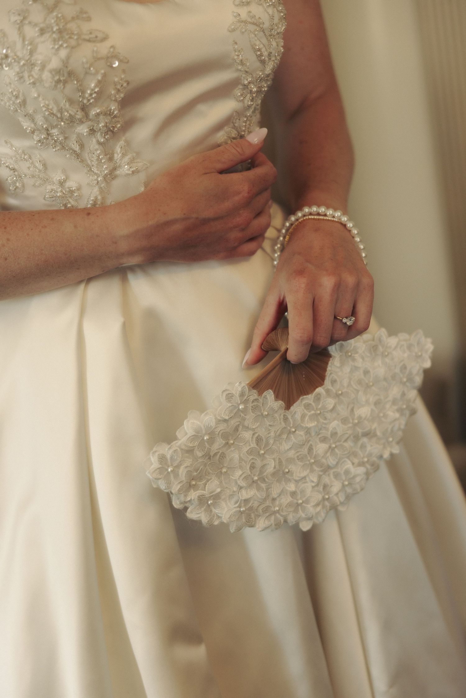 Bride in white lace gown holding decorative floral clutch purse with pearl bracelet and wedding ring.