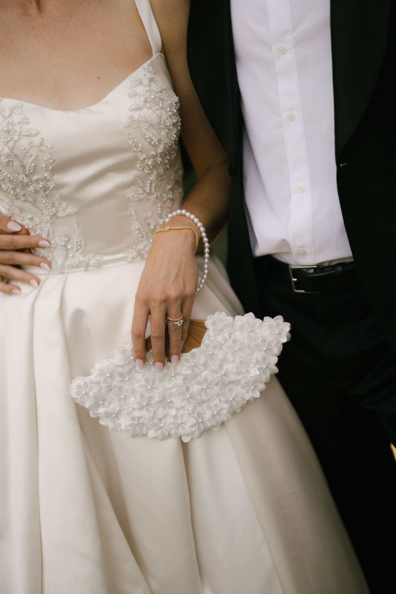 Bride in white lace gown holds floral clutch beside groom in black suit, showcasing wedding ring and pearl bracelet.
