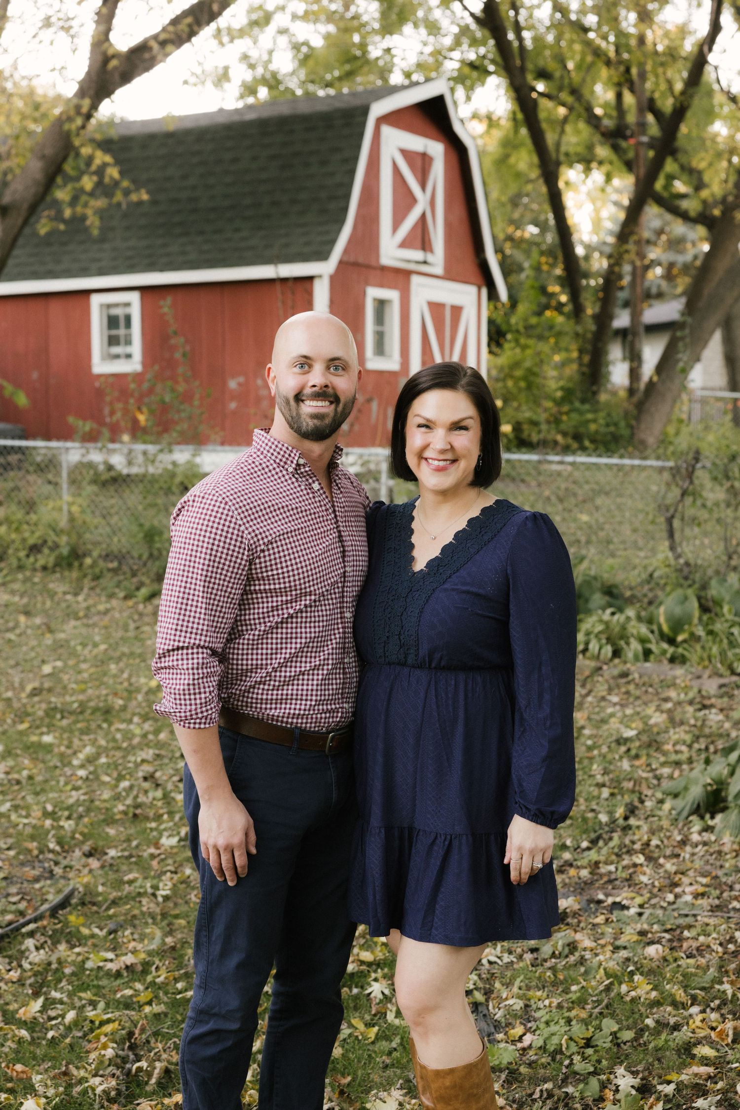 Couple poses together outdoors in autumn with red barn buildings and trees in background.