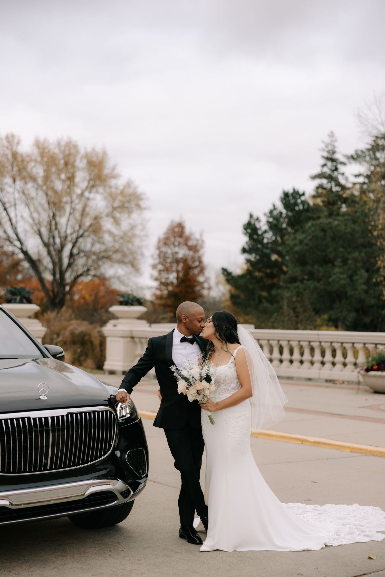 A romantic wedding couple embraces next to a luxury black car on an overcast autumn day.