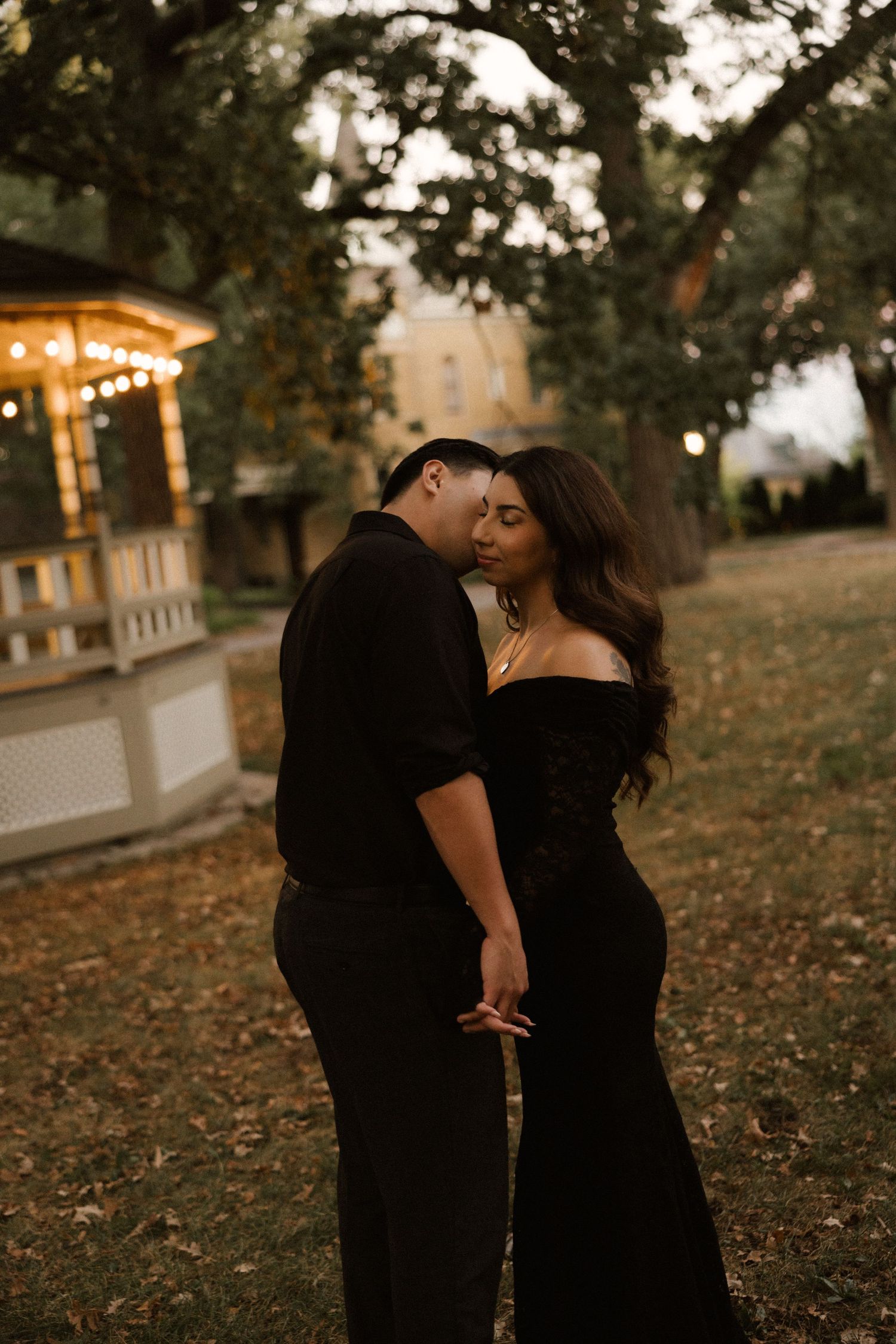 Couple in matching black outfits sharing a romantic moment outdoors near a lit gazebo at dusk.
