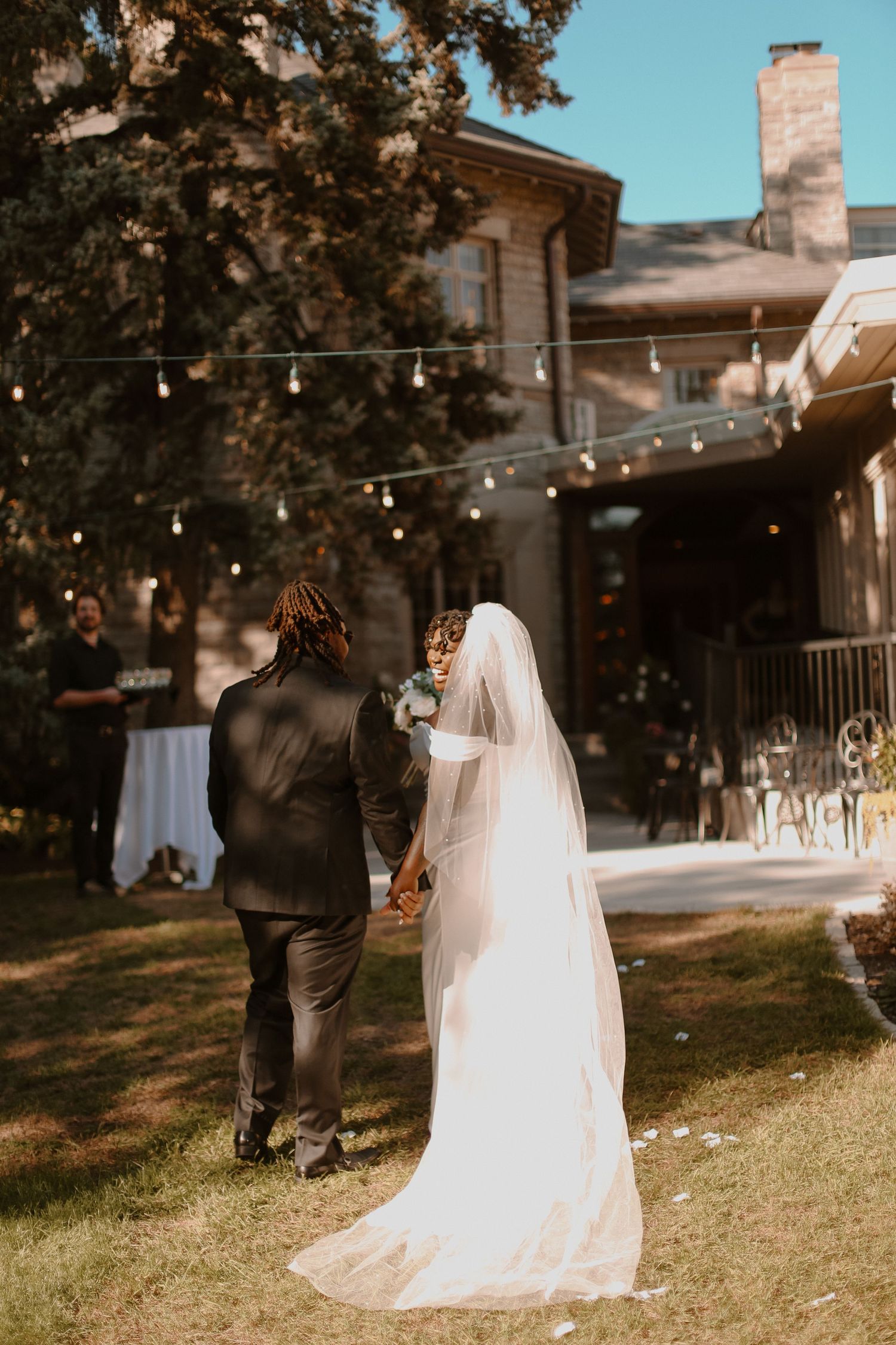 Bride and groom walking towards outdoor reception venue with string lights and rustic decor.