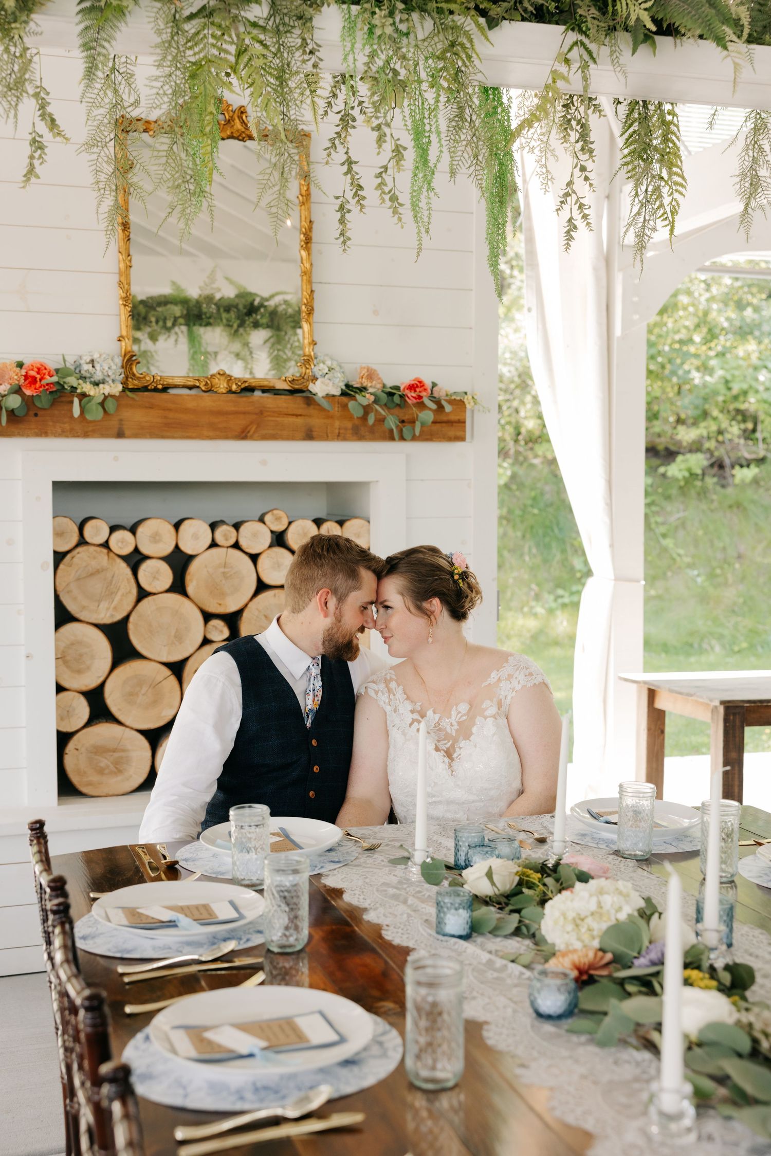 Rustic wedding reception table setting with log wall backdrop, white tablecloths, and elegant place settings.