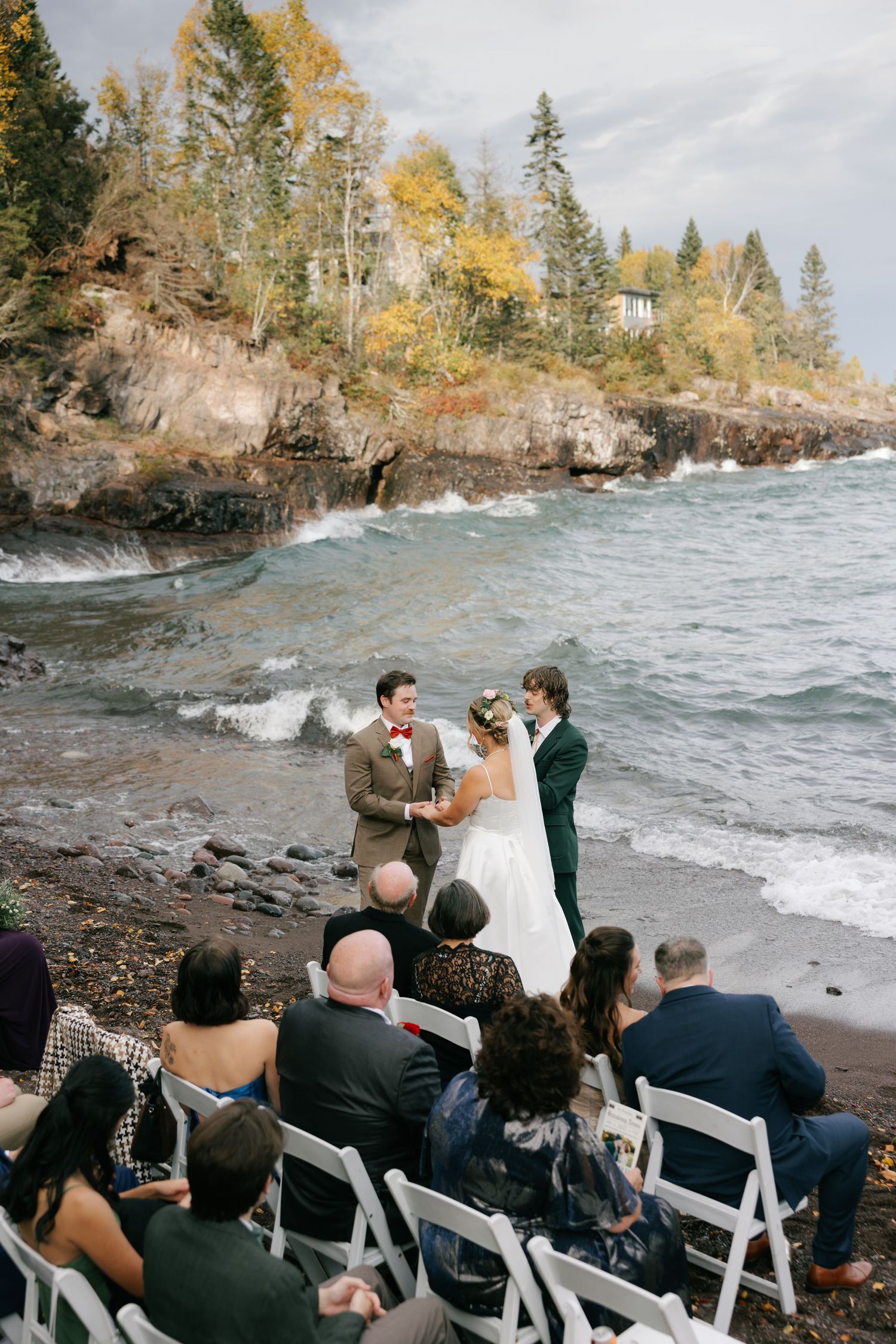 A wedding ceremony takes place on a rocky beach with autumn trees and cliffs along Lake Superior's dramatic shoreline.