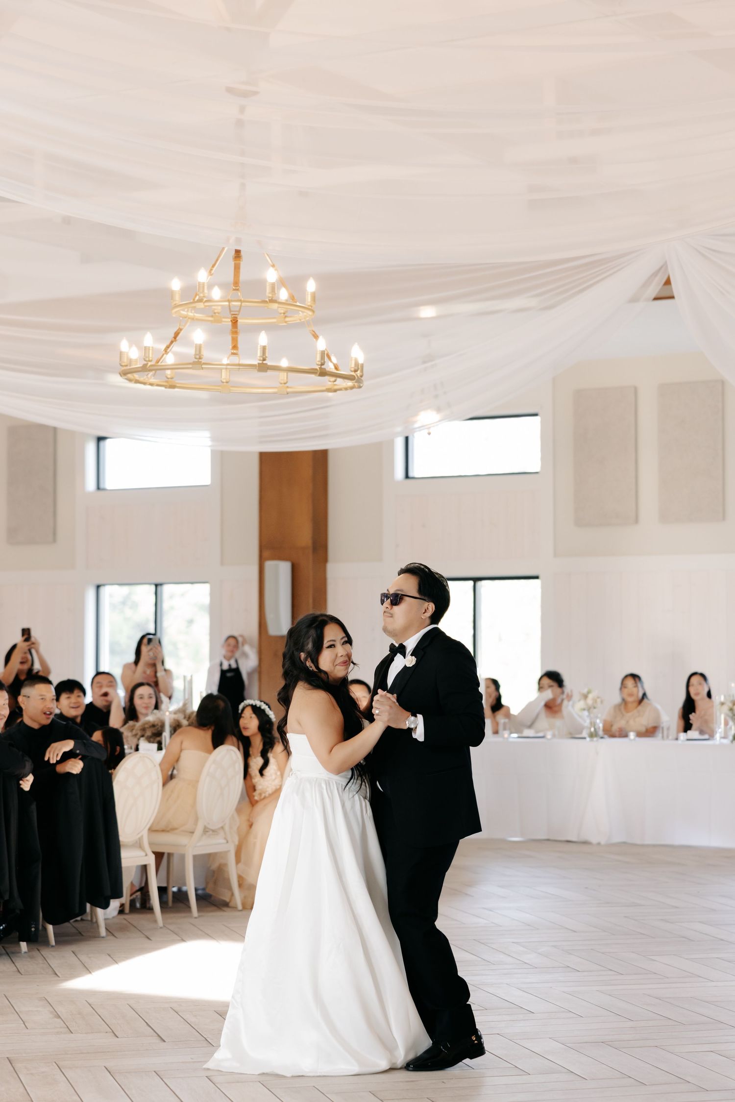 A couple shares their first dance at a wedding reception in an elegant white room with draped ceiling and chandelier.