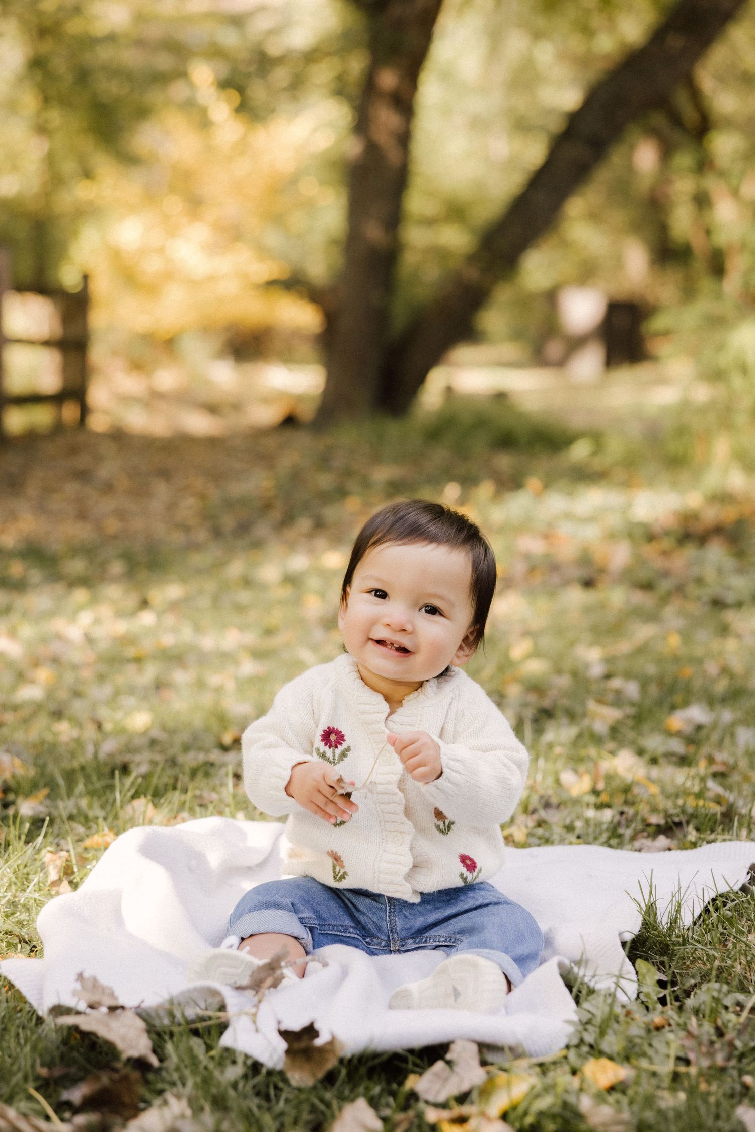 A baby sits on a white blanket outdoors in autumn, wearing a white embroidered top and jeans while smiling at the camera.