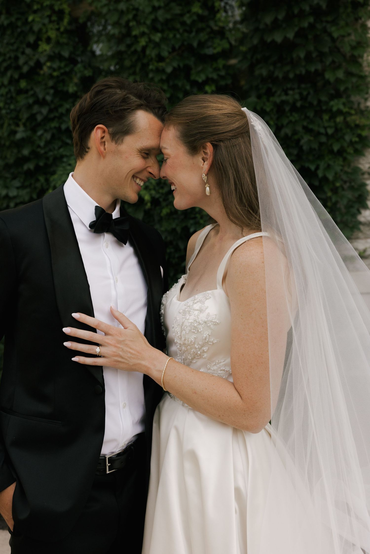 Bride and groom smile forehead-to-forehead, she in a white gown with veil, he in a black tuxedo, outdoors.