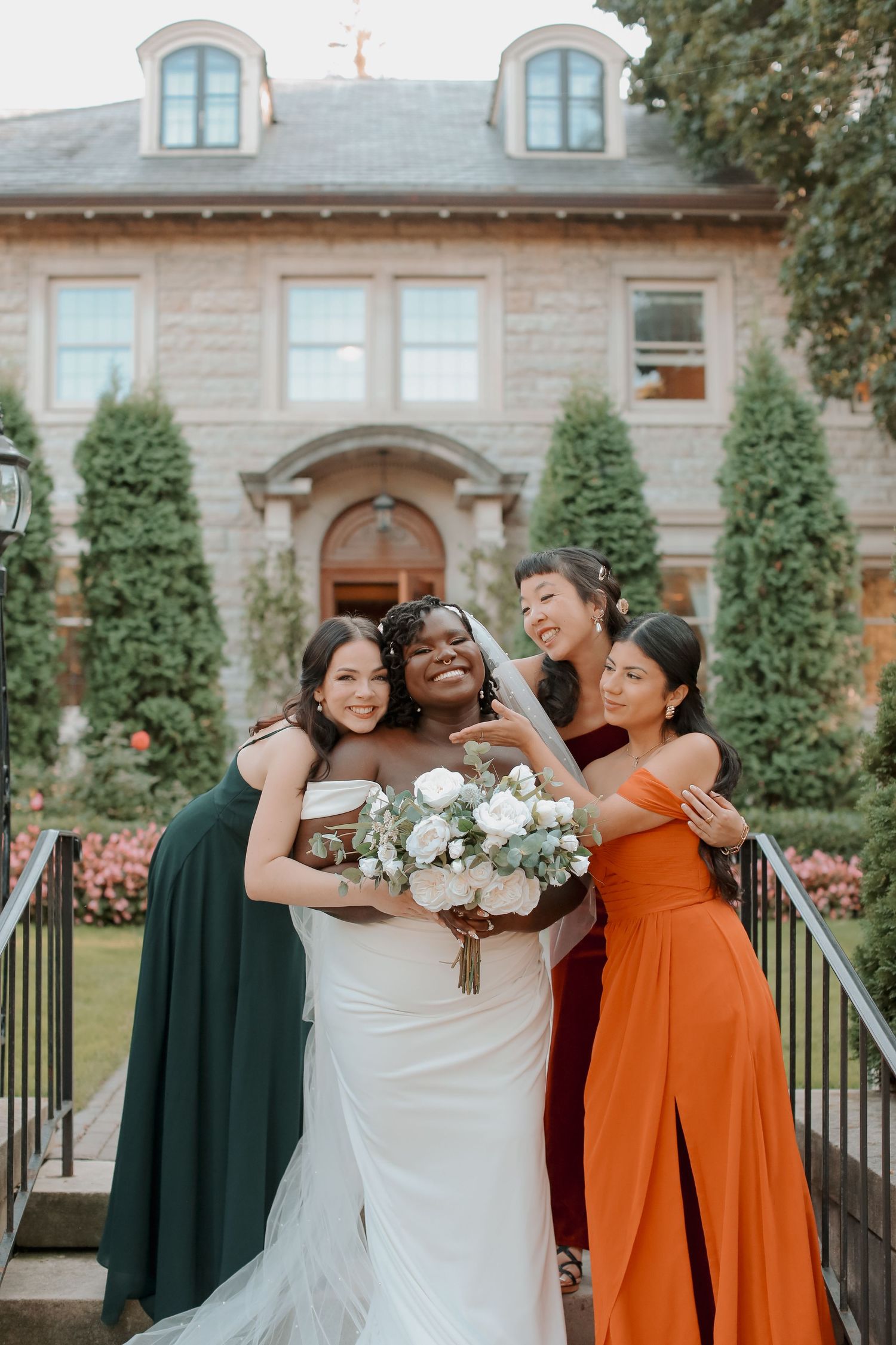 Bride surrounded by bridesmaids in orange and green dresses share joyful moment.