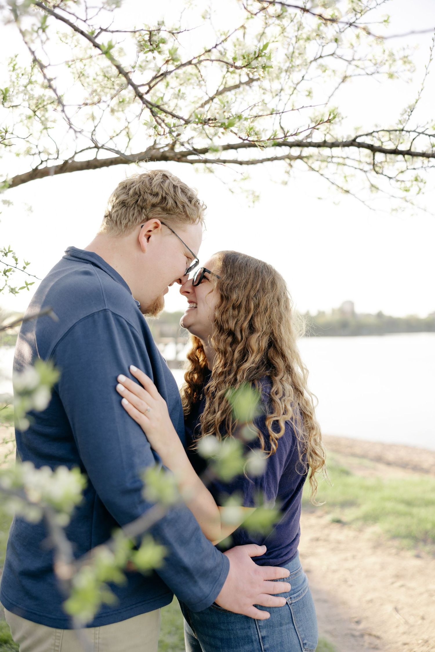A romantic couple in navy blue embraces under blooming spring tree branches beside a tranquil lake.