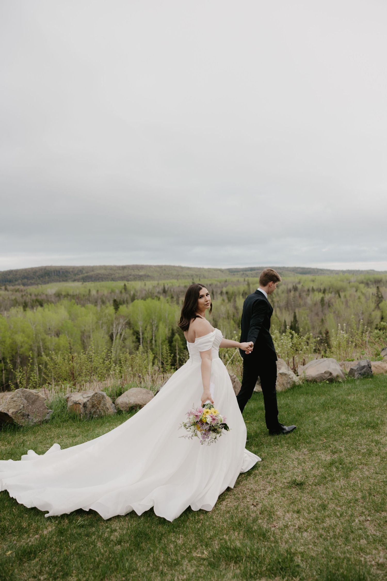 A couple stands on a hilltop overlooking rolling green hills, with the bride's white flowing dress trailing behind on the grass.