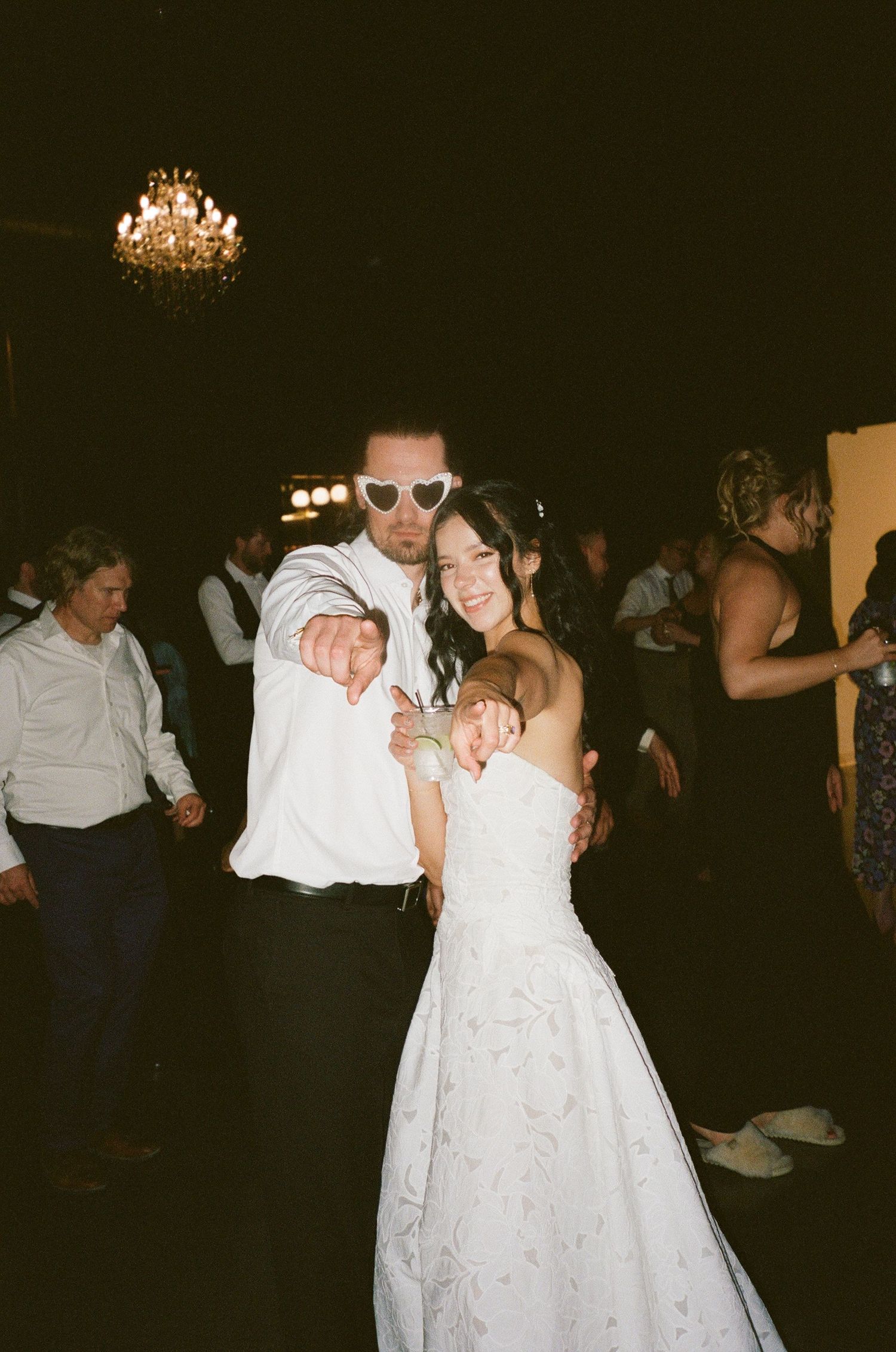 Bride in white gown and groom in white shirt with heart sunglasses dancing at wedding reception under chandelier.