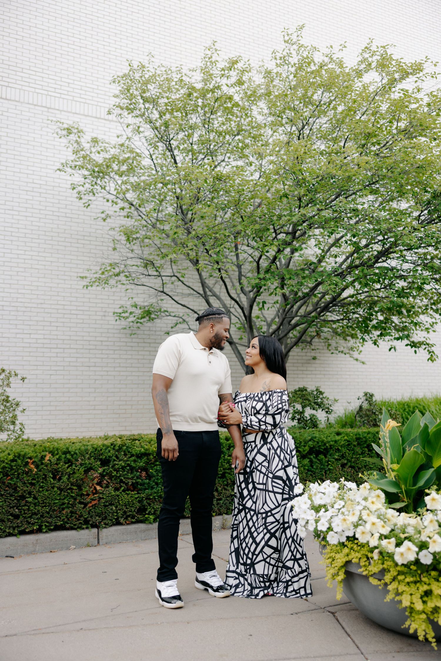 Couple sharing a romantic moment in a garden with blooming white hydrangeas against a white brick wall.