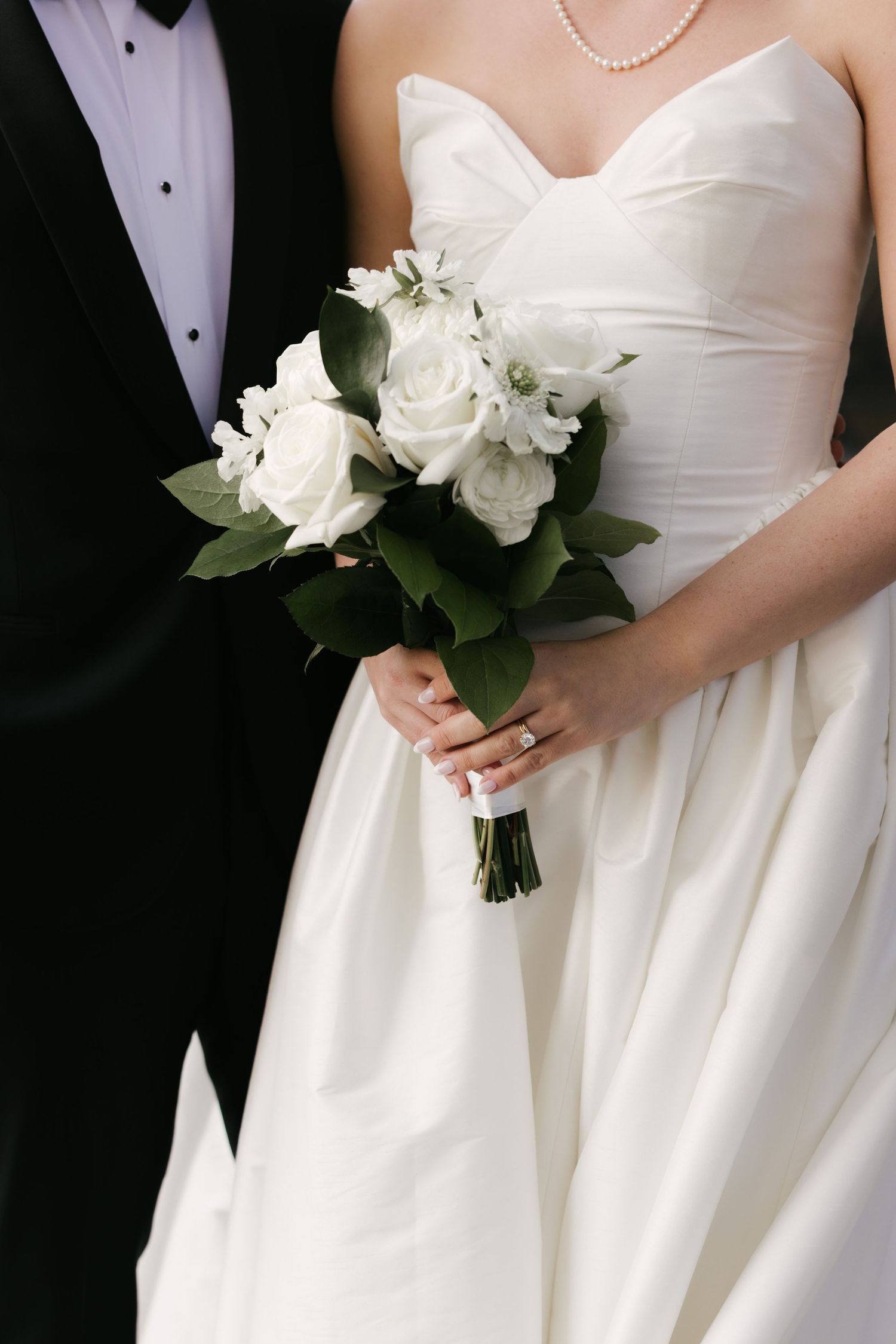 Bride in strapless white gown holds elegant bouquet of white roses and greenery beside groom in black tuxedo.
