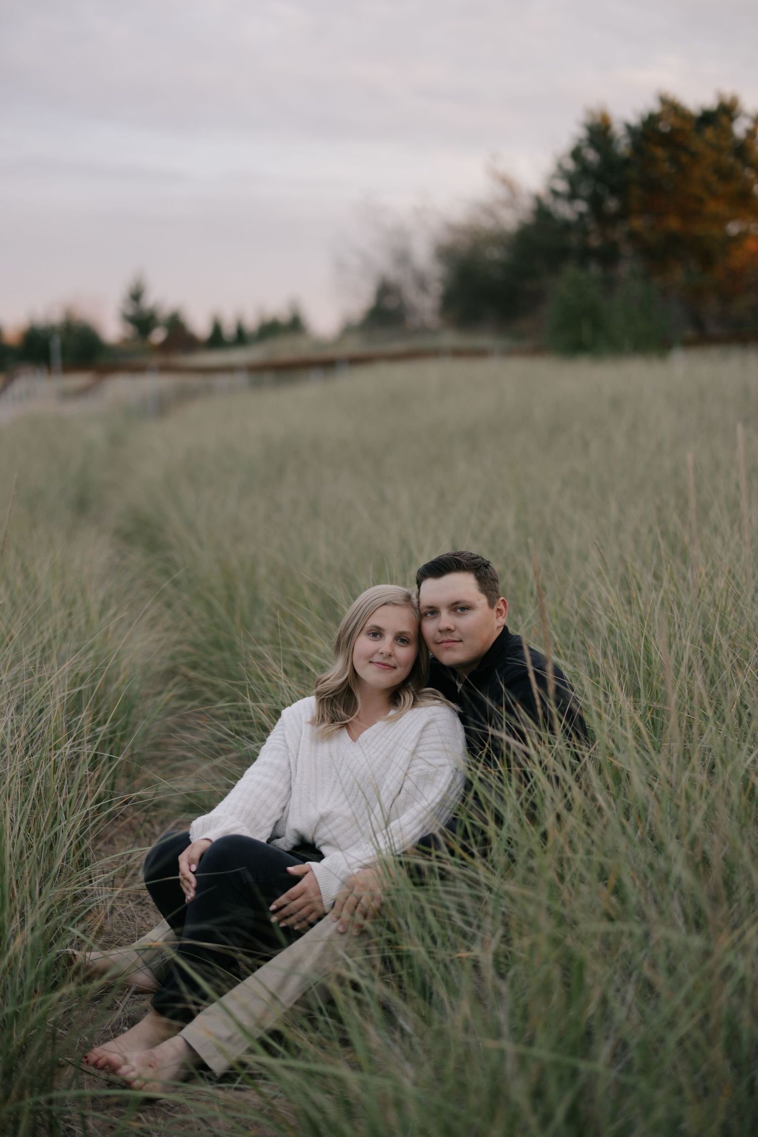 A couple sits together in tall beach grass during sunset at a coastal location.