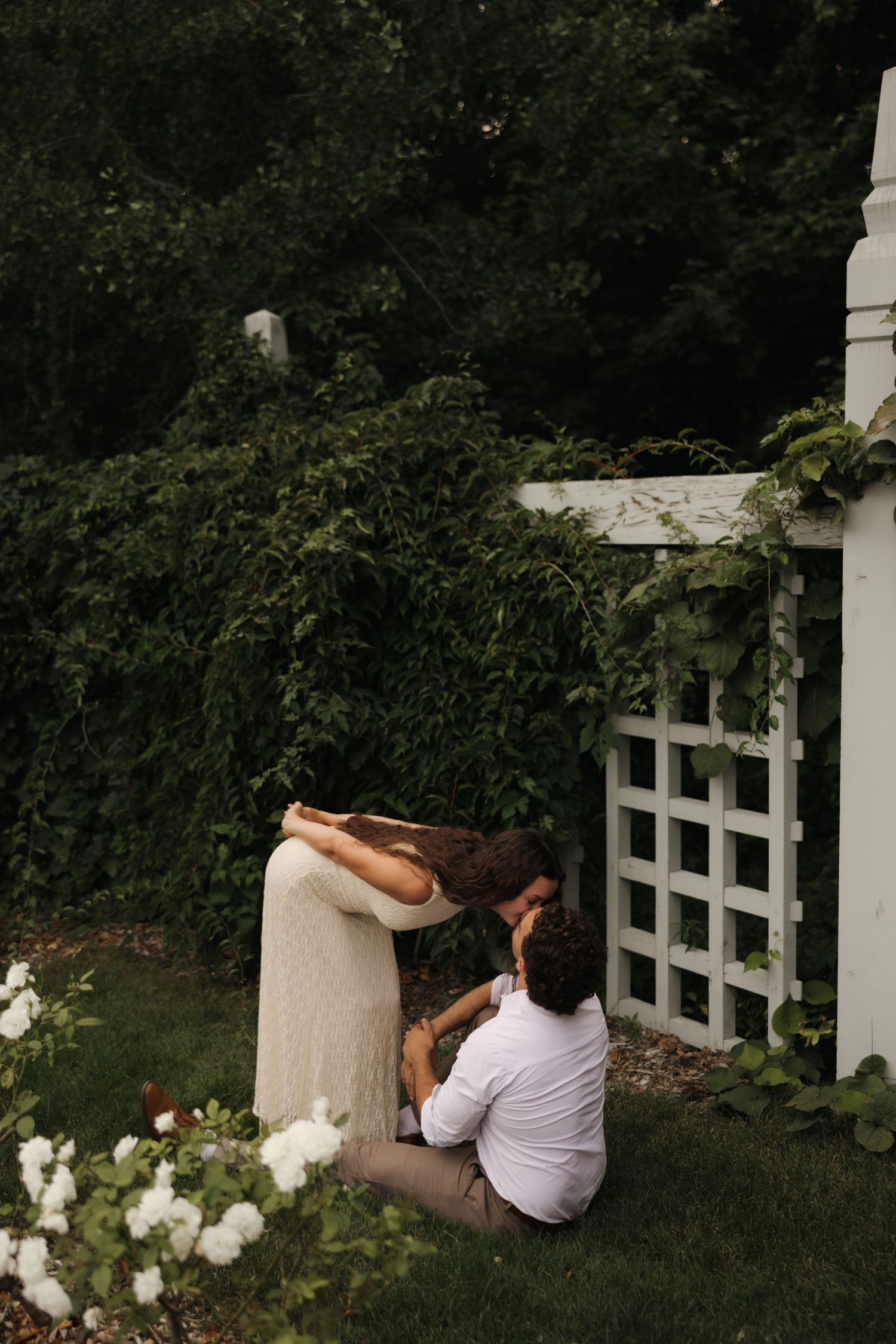 Couple sharing intimate moment in lush garden beside white lattice trellis surrounded by greenery and white flowers.