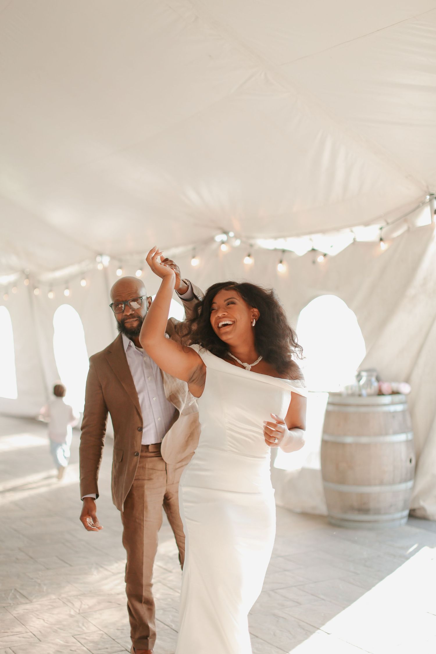 Happy couple dancing and celebrating under a white tent with string lights and wine barrels.