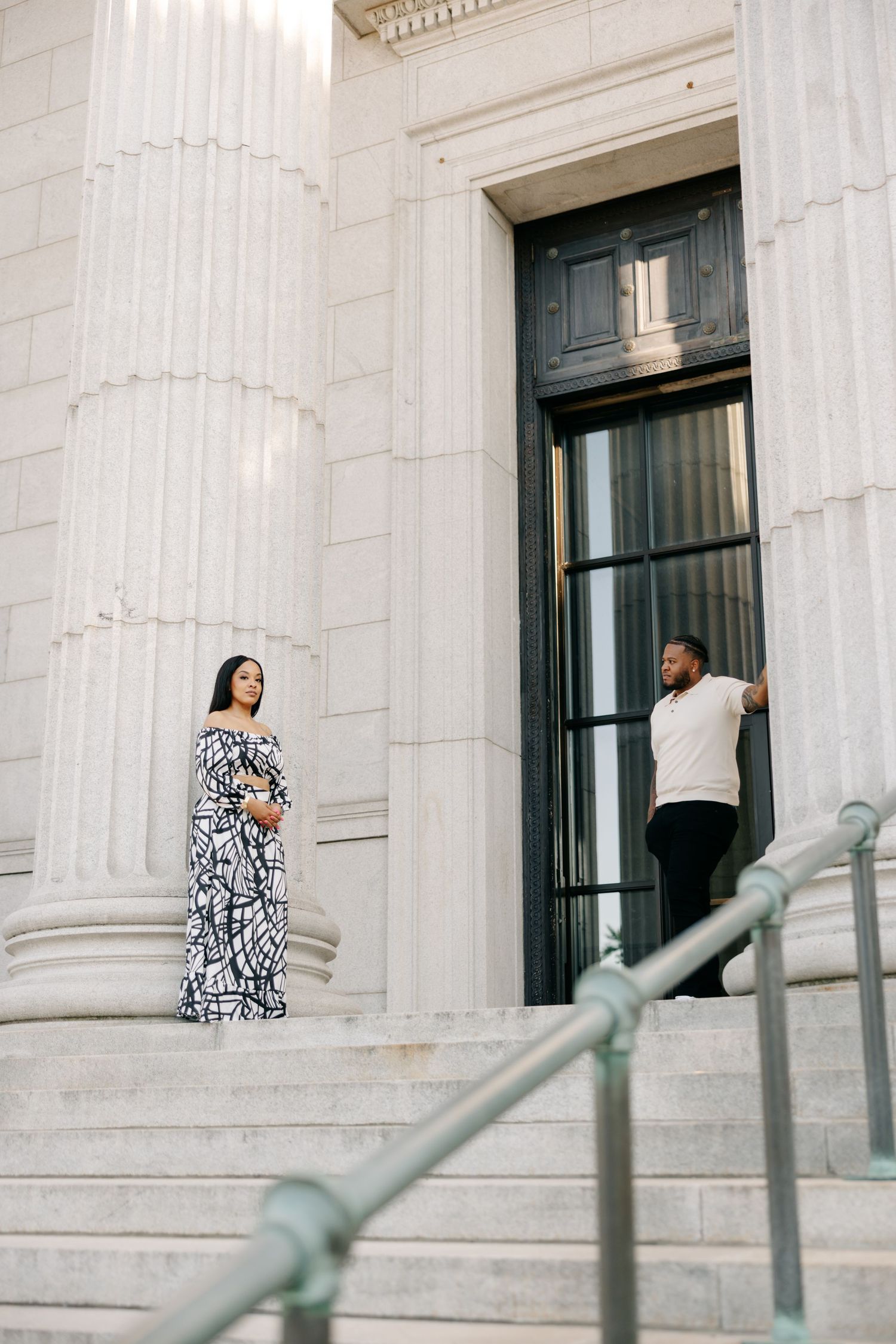 Pregnant woman in black and white maxi dress poses on marble steps with man near ornate building columns.