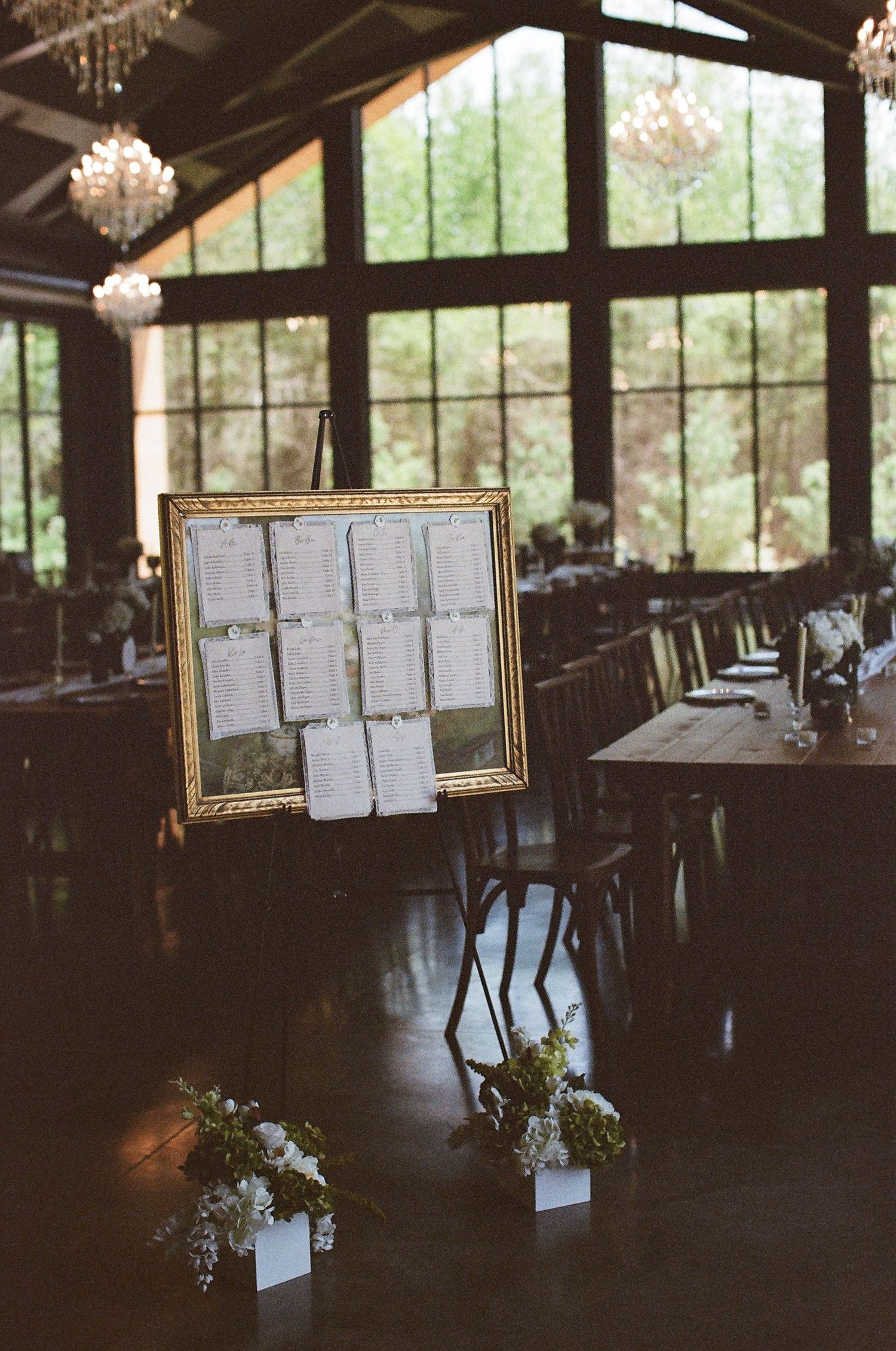Elegant wedding reception seating chart in gold frame on easel with floral arrangements and chandelier-lit venue.