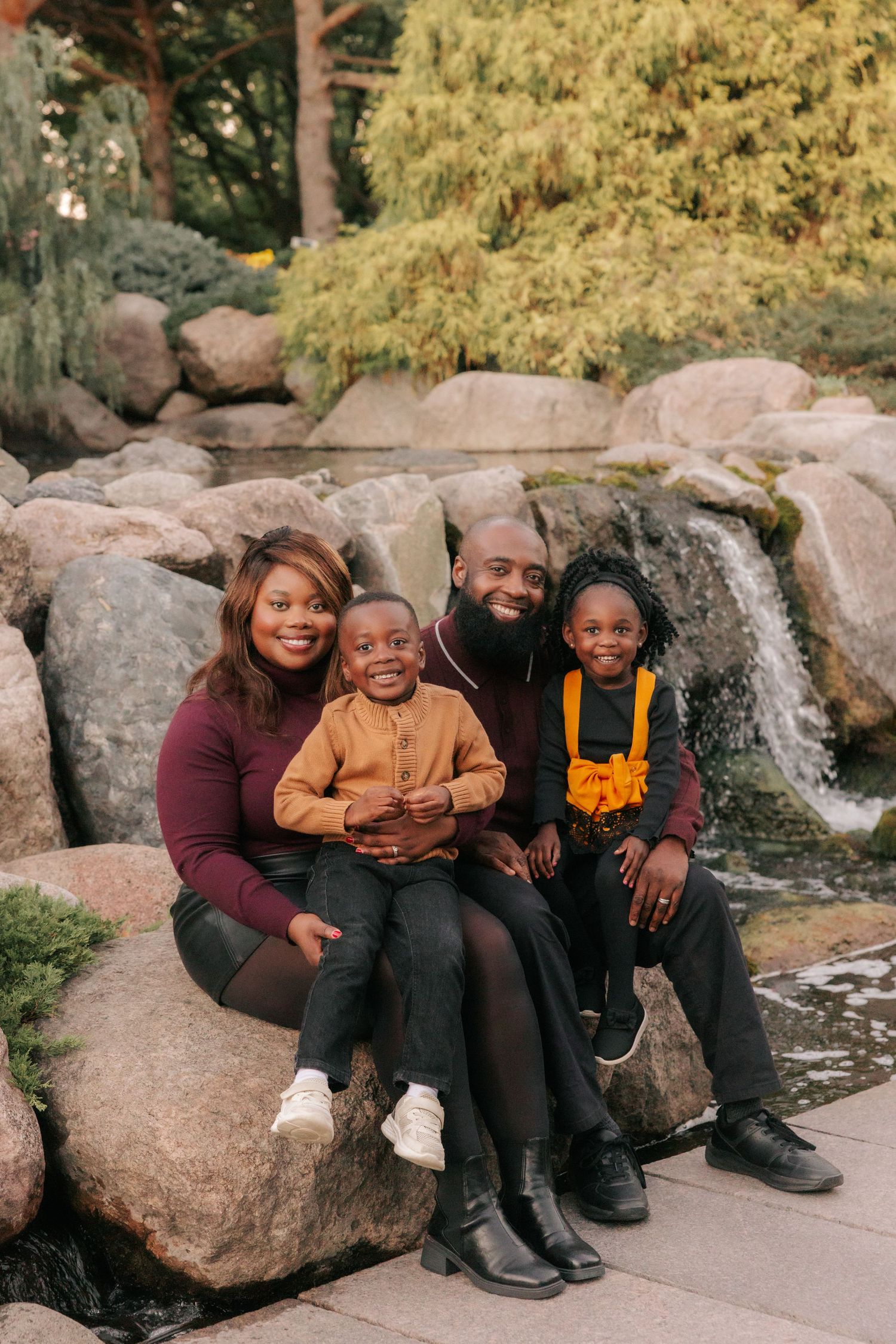 A family sits together on large rocks by a waterfall in an autumn park setting.