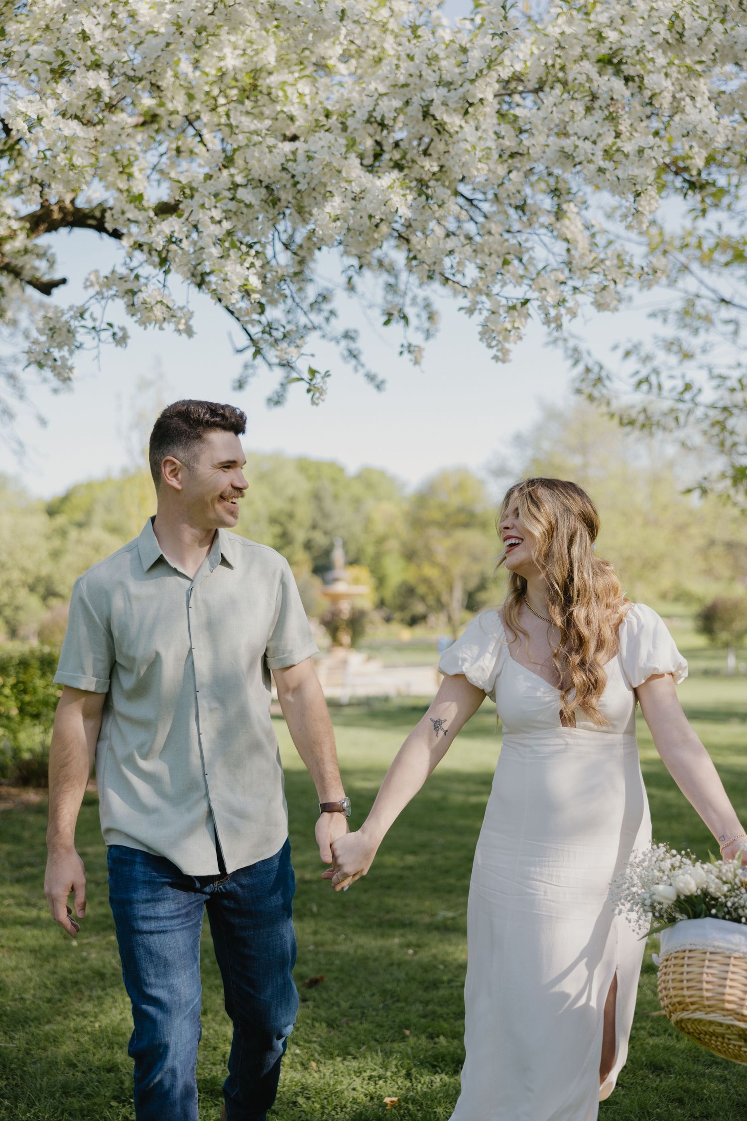 A couple holding hands walking under blooming white cherry blossoms in a spring garden setting.