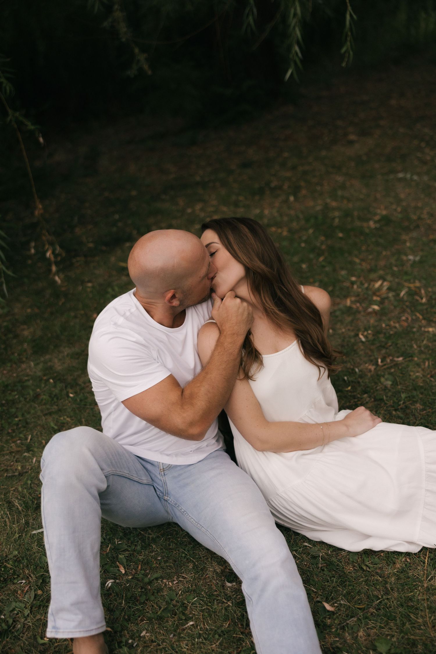 A couple in white outfits share a romantic kiss while sitting on green grass beneath weeping willow trees.