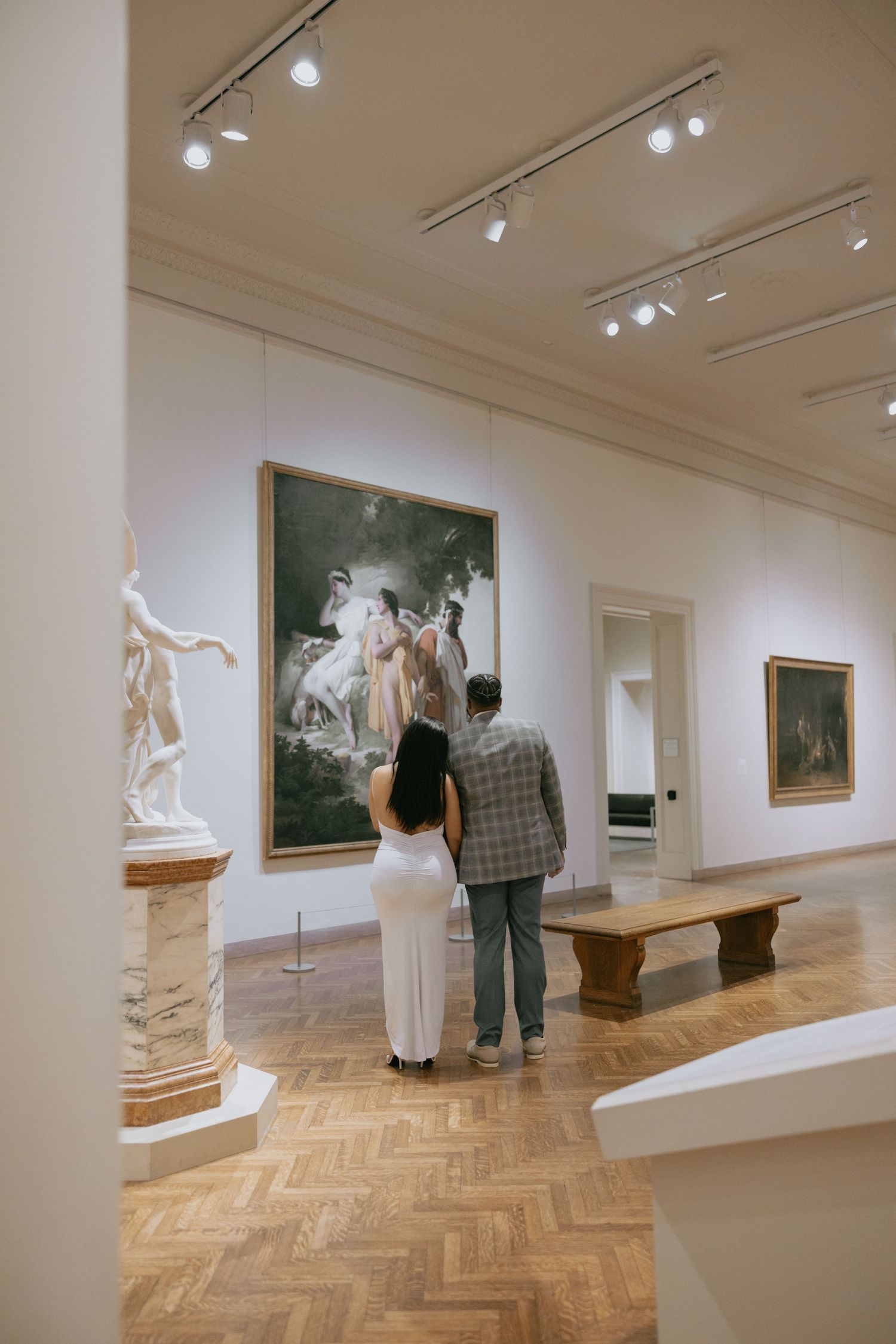 Gallery visitors view artwork in a museum with herringbone wood floors and track lighting illuminating white walls.
