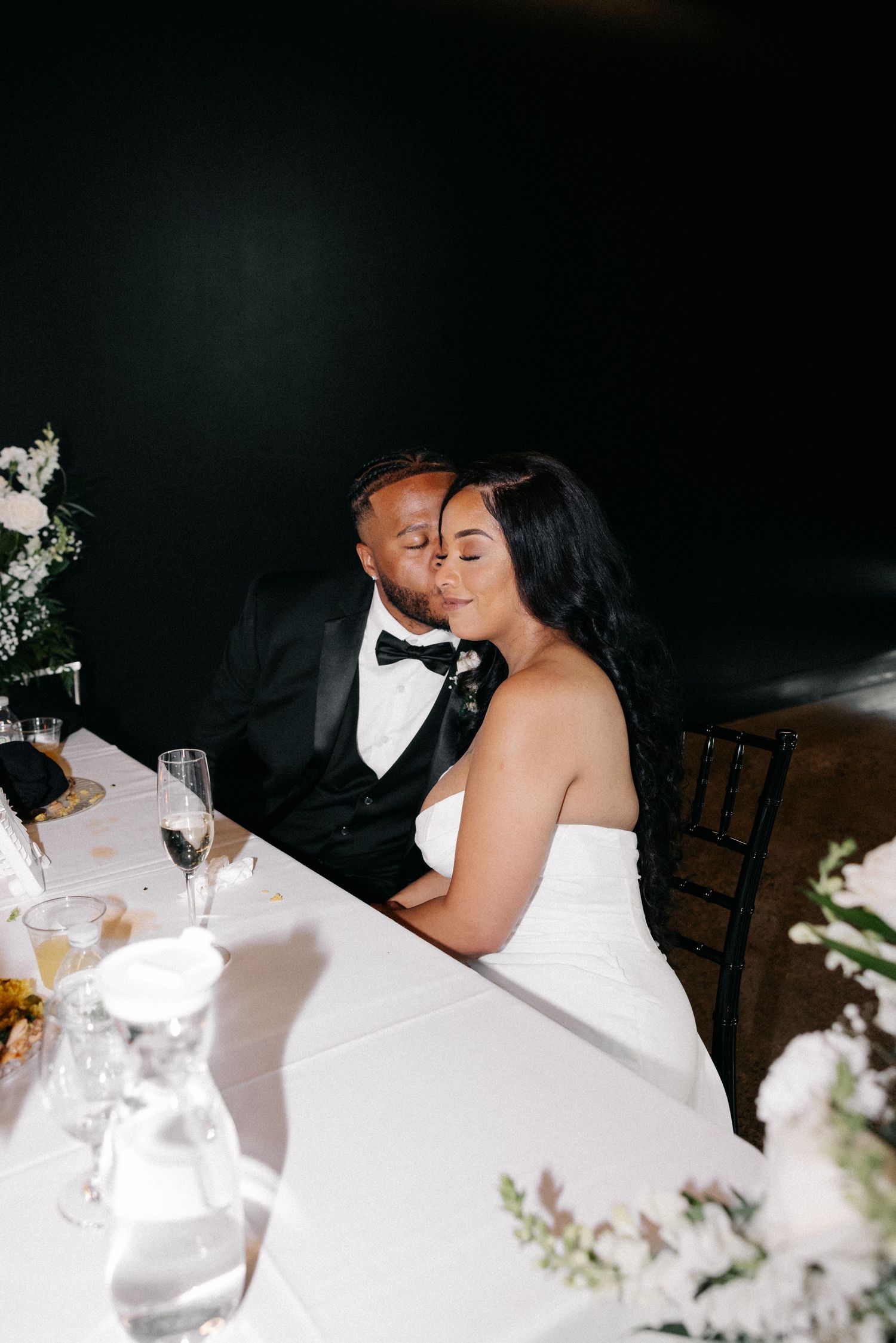 A romantic wedding reception moment showing a couple sharing a kiss at their table decorated with white floral arrangements.