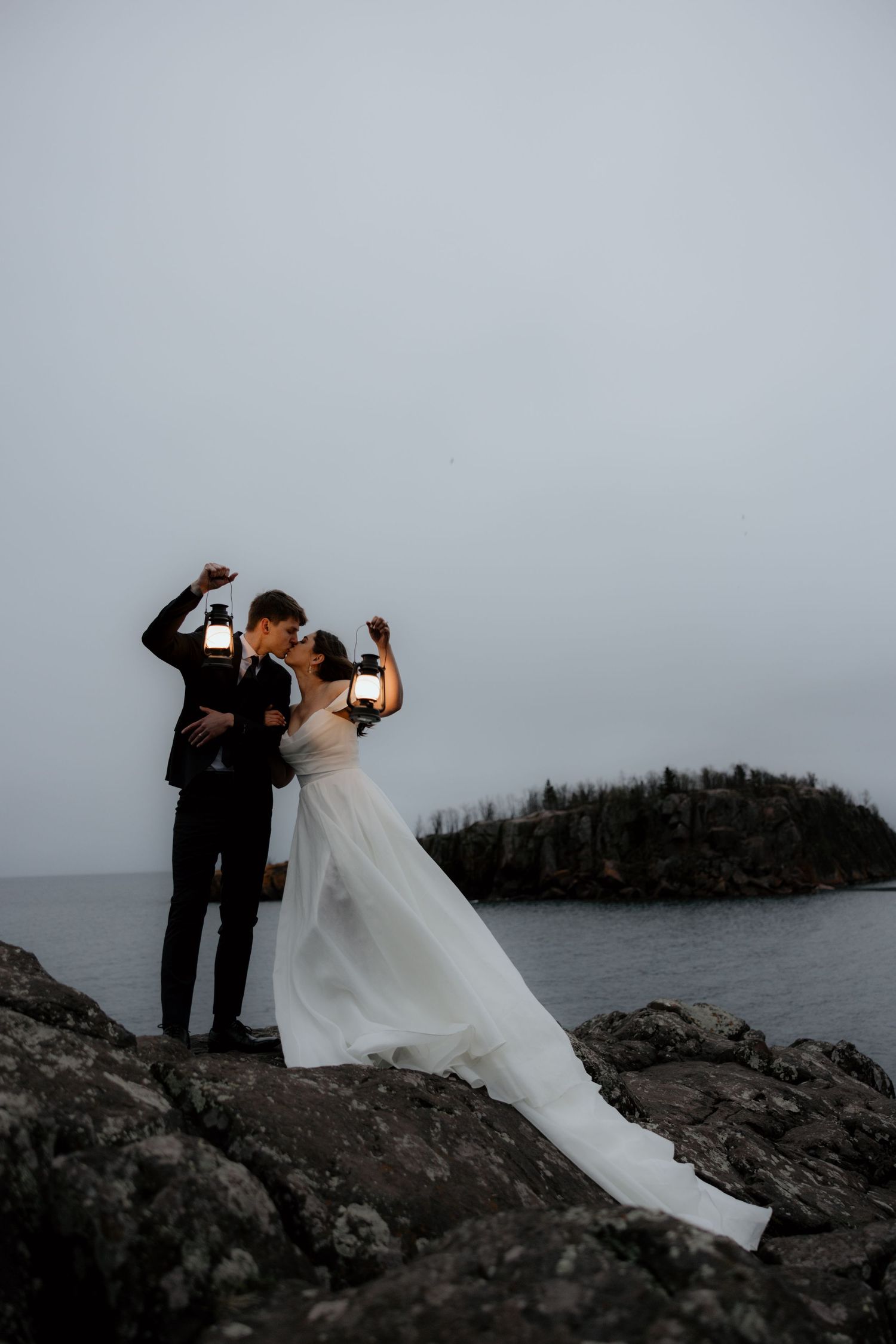 Wedding couple holding lantern stands on rocky coastal cliff overlooking island at dusk.