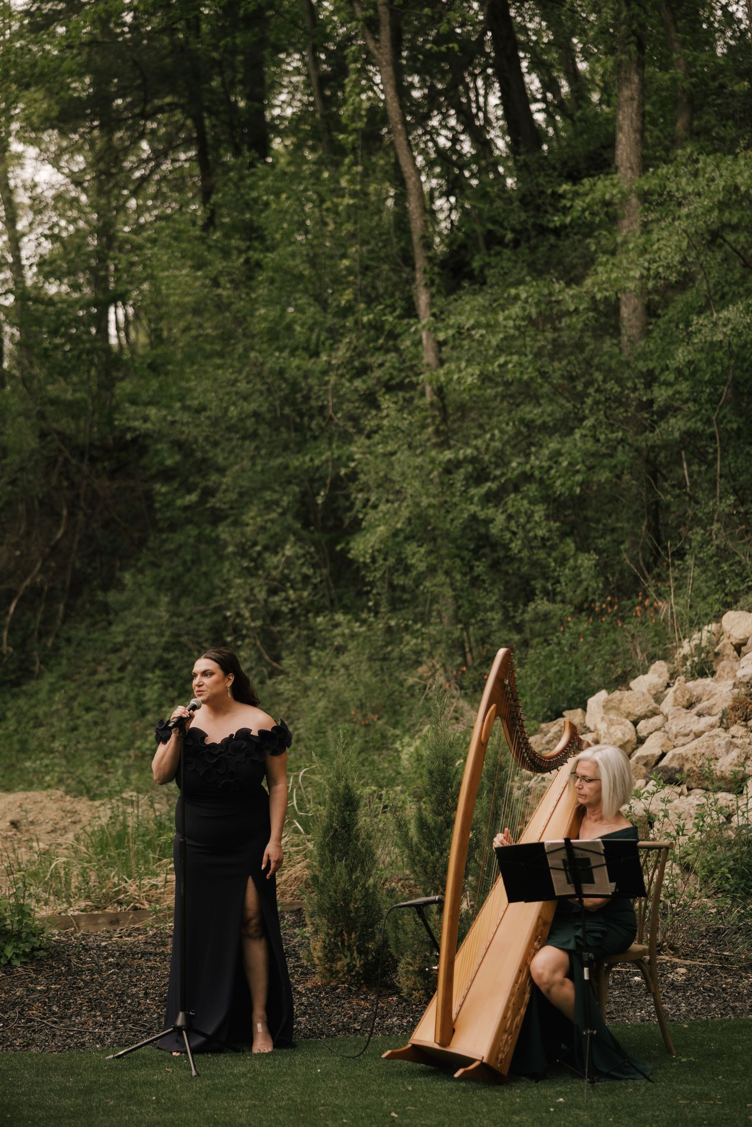 A vocalist in a black gown performs beside a harpist outdoors at an elegant wedding ceremony.