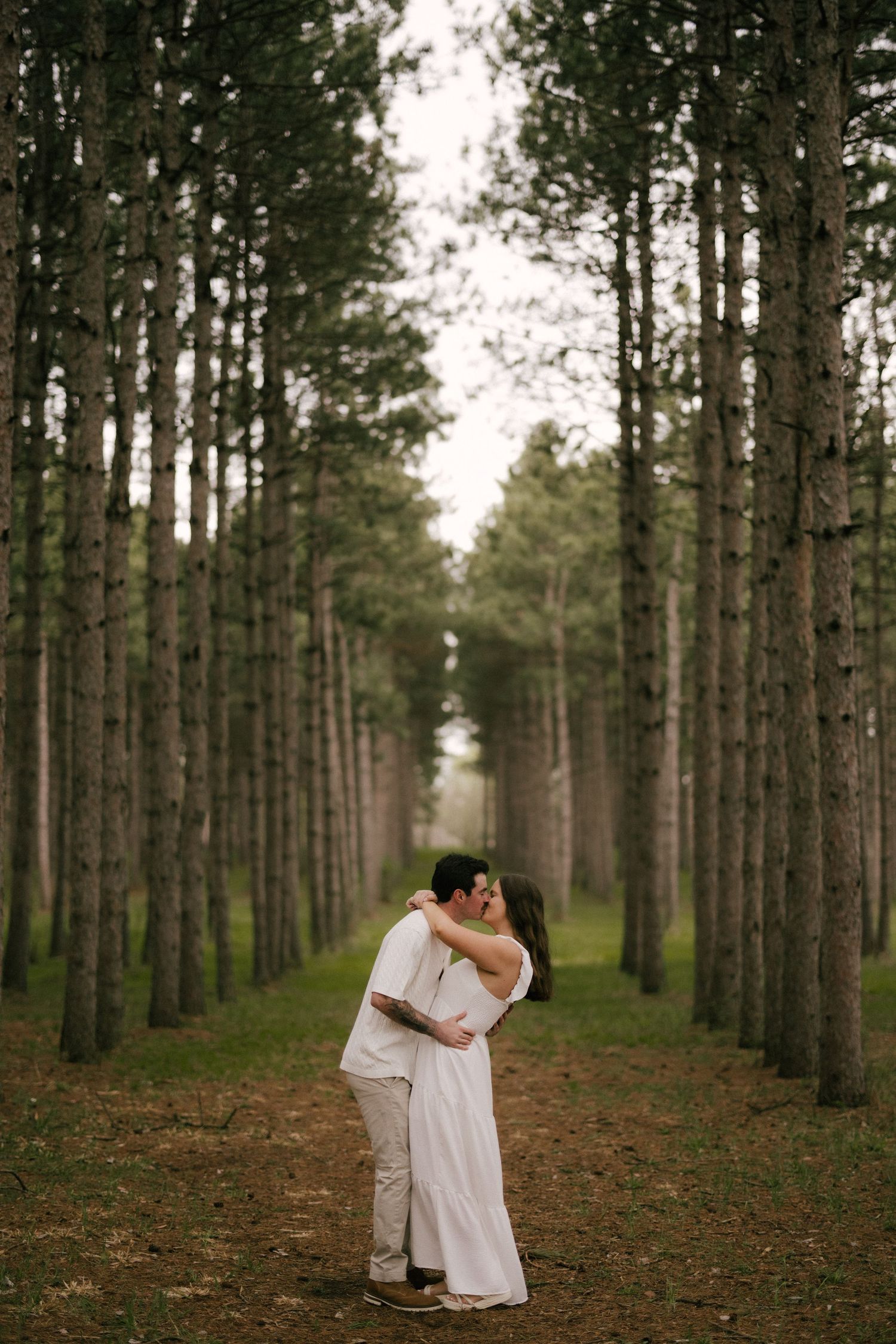 A couple shares a romantic kiss among tall pine trees in a lush forest, dressed in elegant white attire.