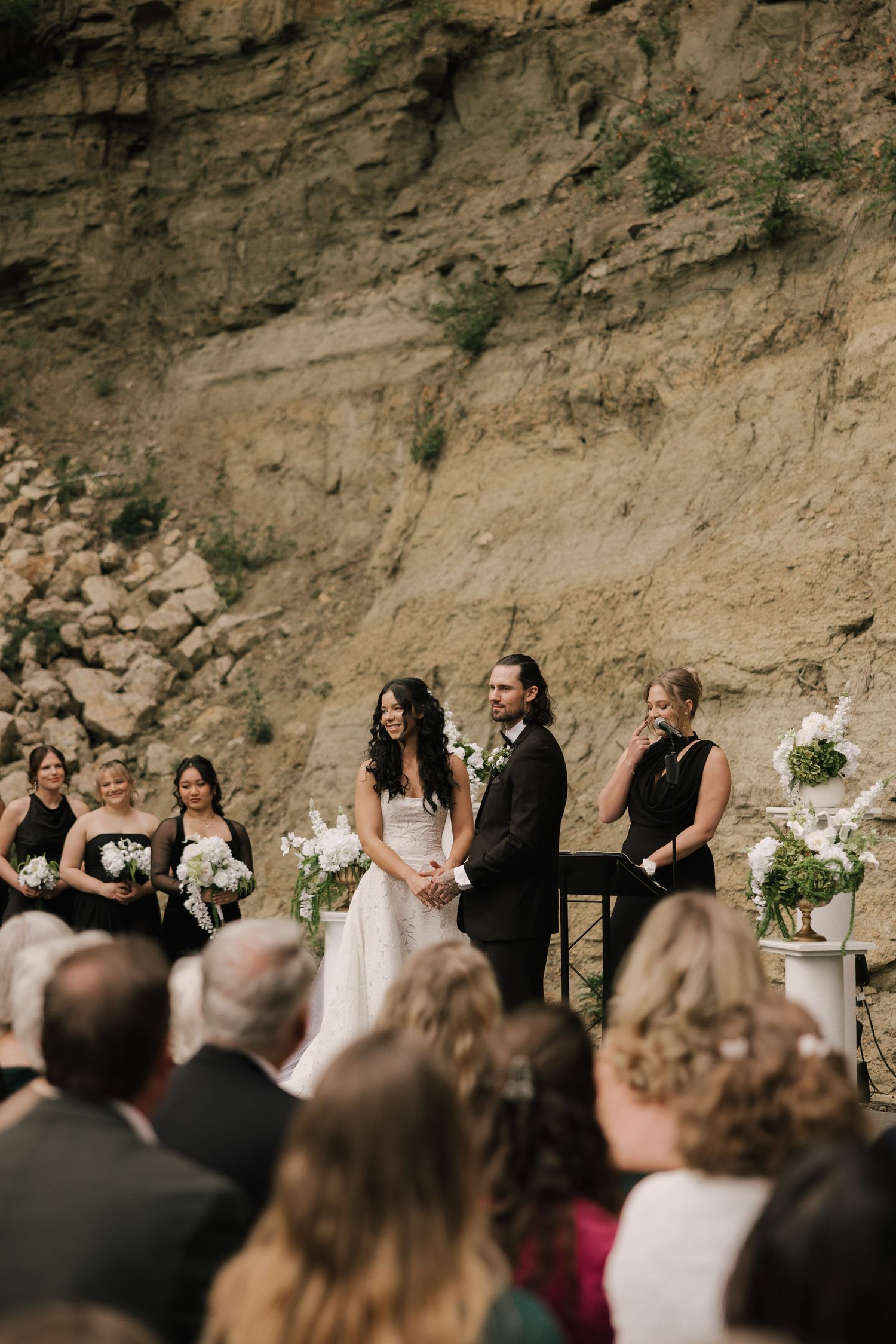 Bride and groom hold hands at outdoor wedding ceremony against rocky cliff backdrop with bridesmaids in black dresses.