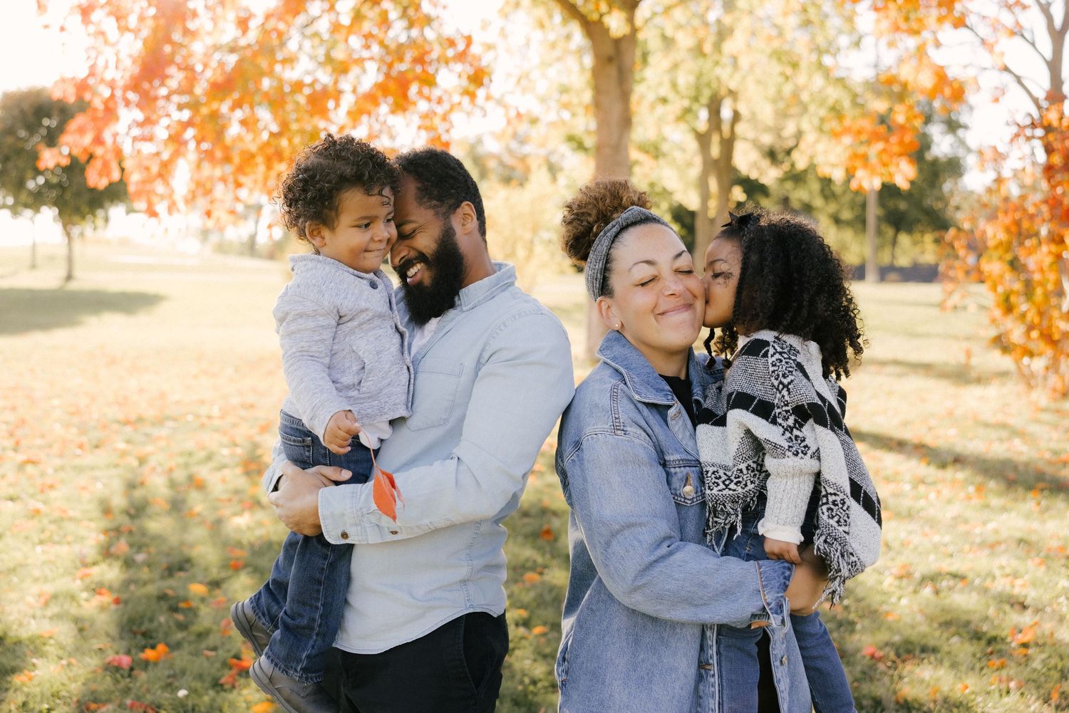 A family shares tender moments together in a park during autumn with golden leaves in the background.