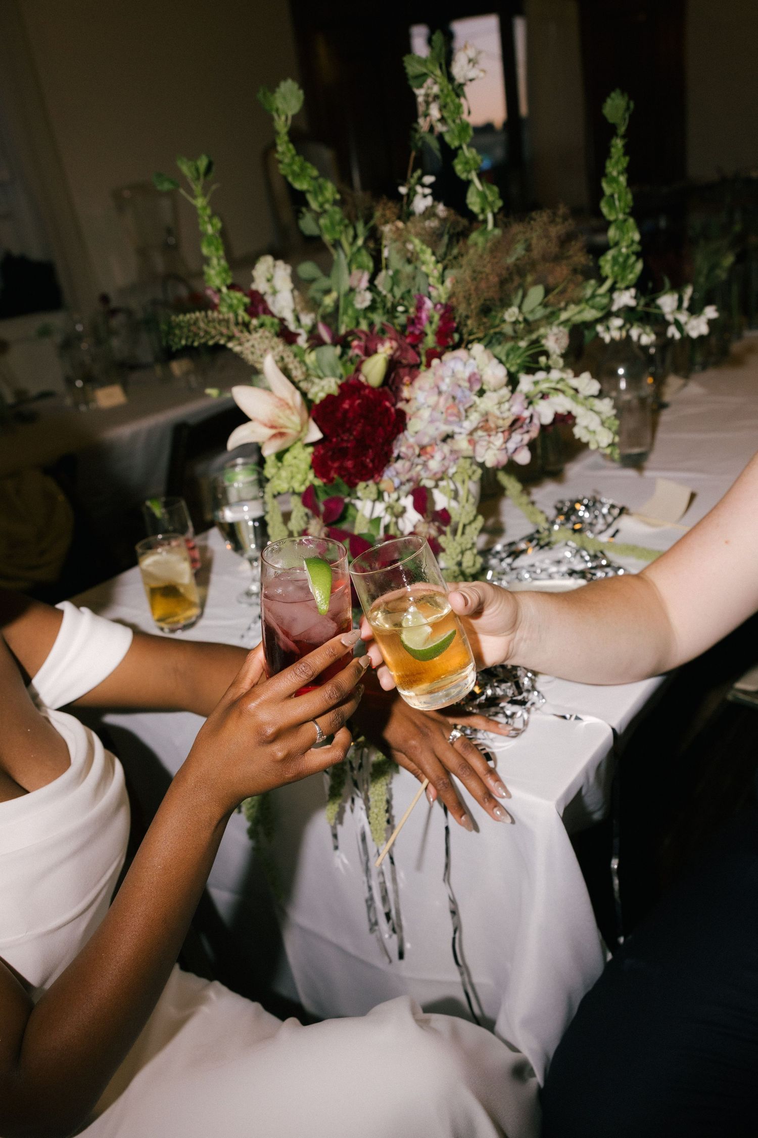 Two women clinking cocktail glasses at an elegant wedding reception table adorned with lush floral centerpiece.