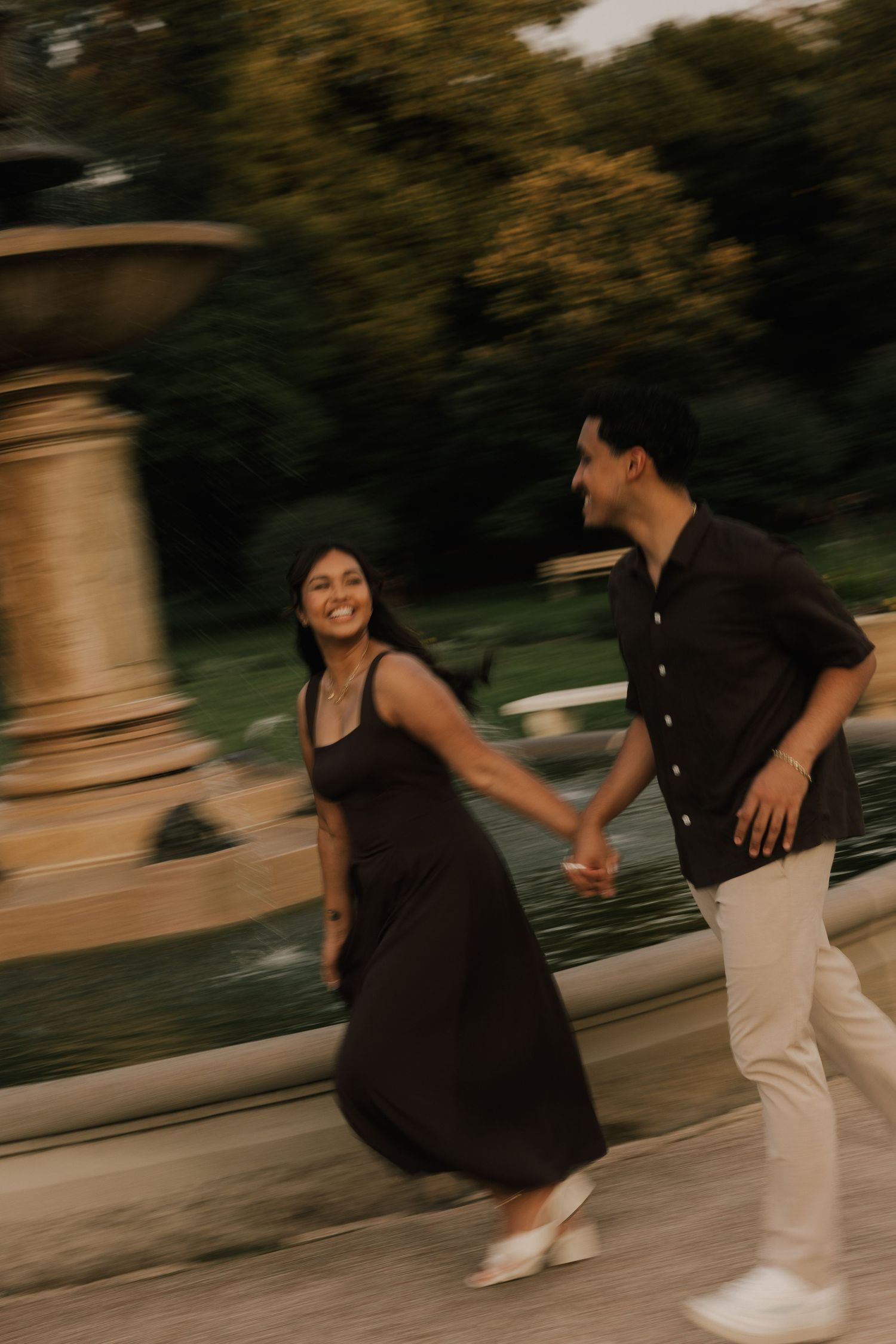 A joyful couple holds hands and walks near a fountain in a park, captured with a motion blur effect.