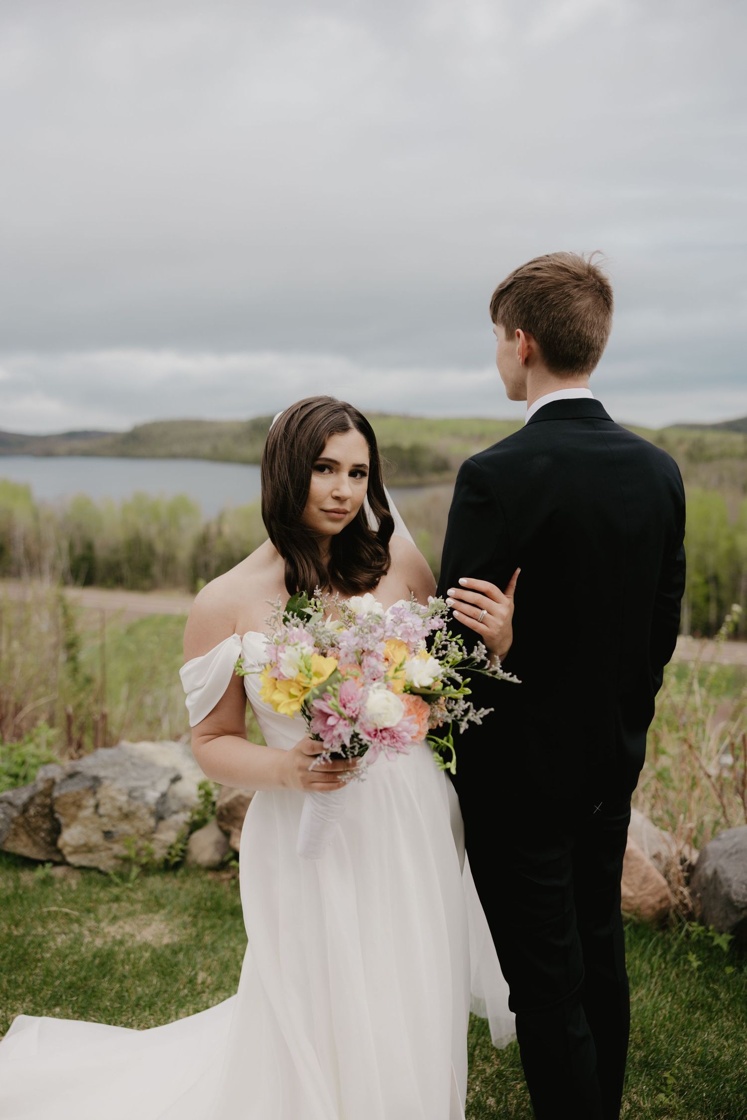 A bride in an off-shoulder white dress holds a pastel bouquet while posing with groom outdoors near a lake.