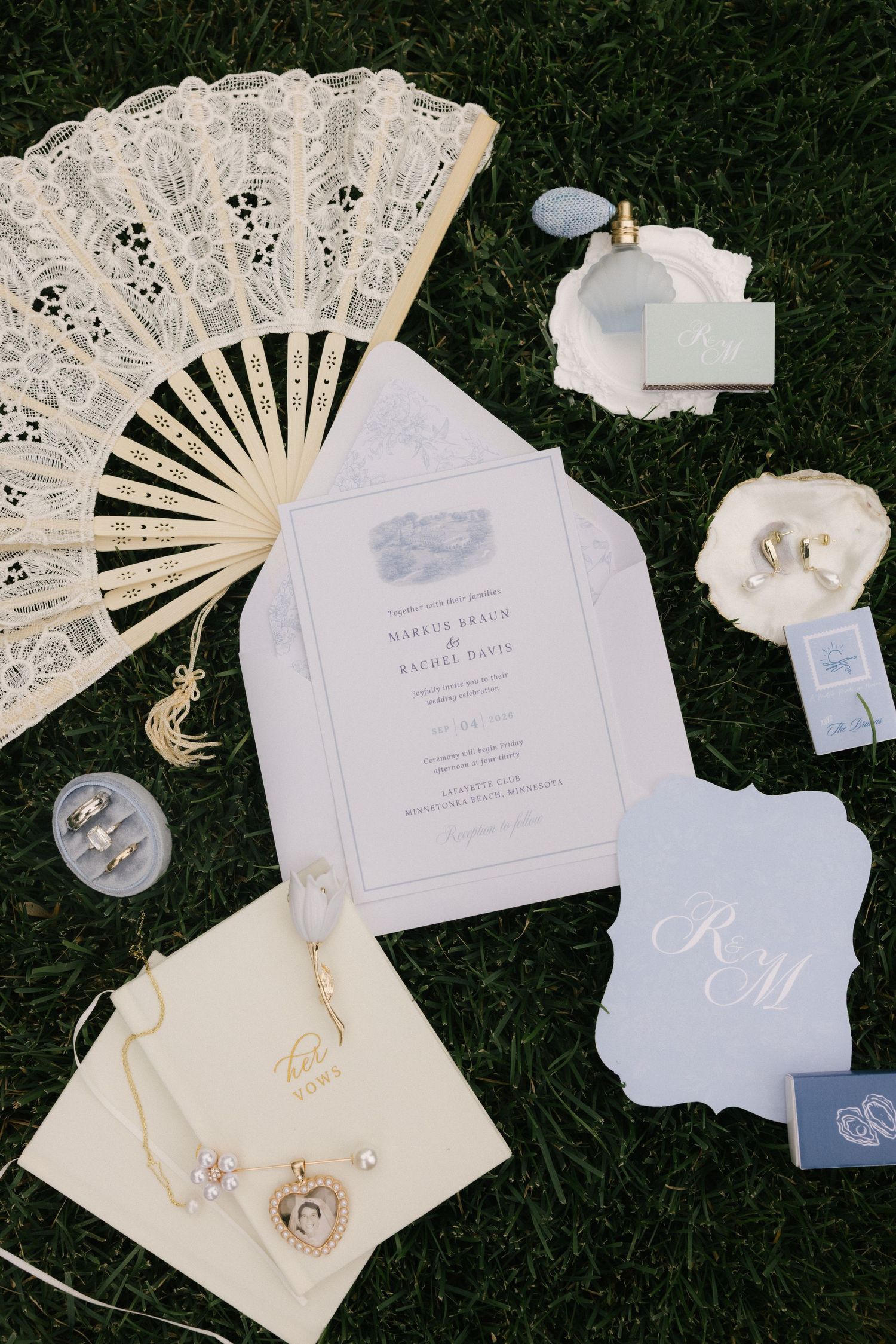 Elegant wedding flat lay with lace fan, floral invitations, rings, perfume, and monogram stationery on green grass.