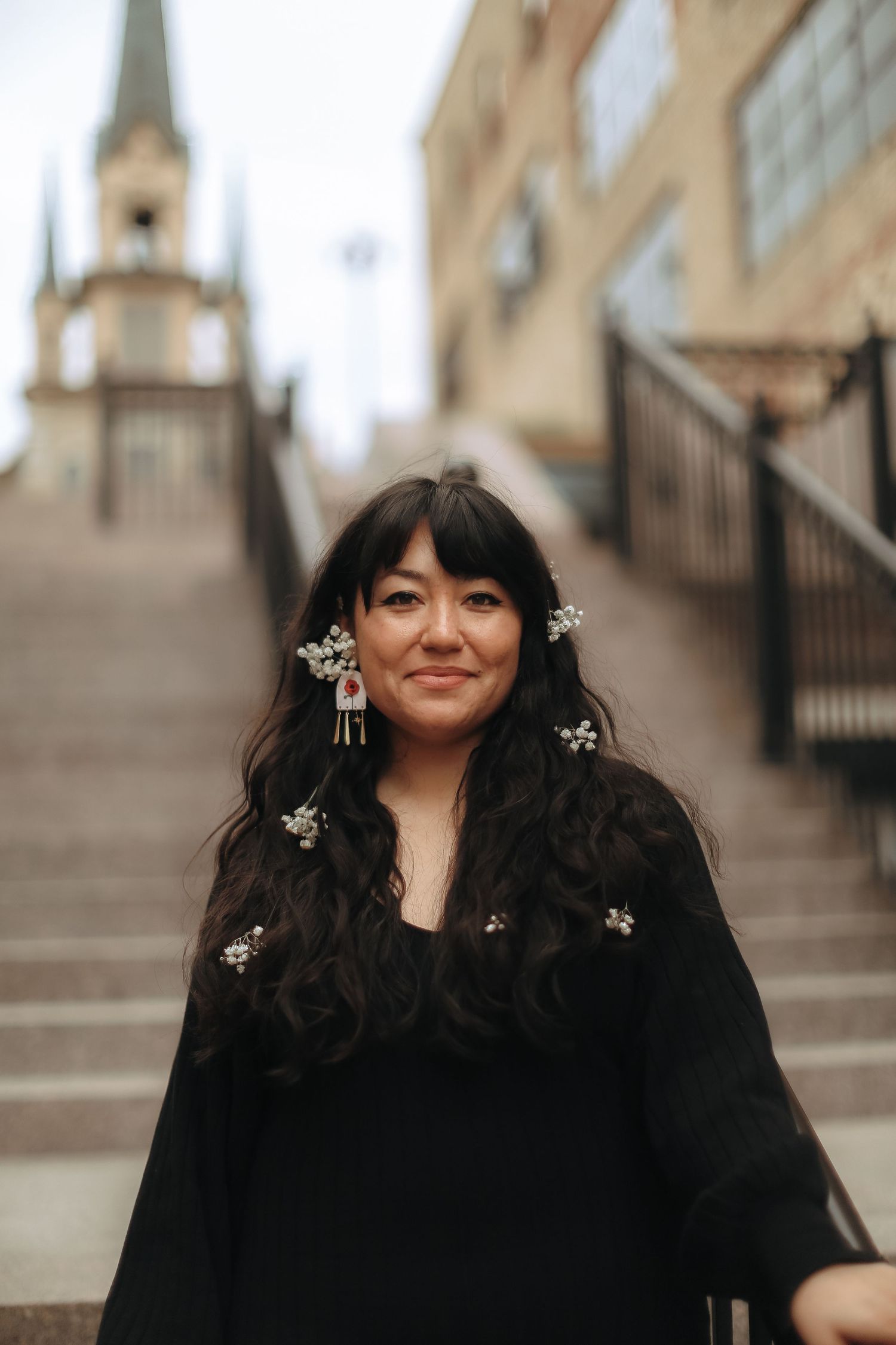 A person with small flowers in their dark hair stands on stone steps near a church spire.