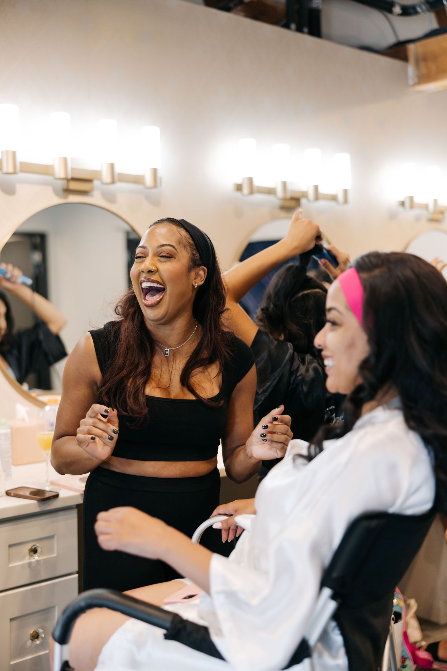 Two friends laughing together while sitting at a makeup vanity in a bright room.