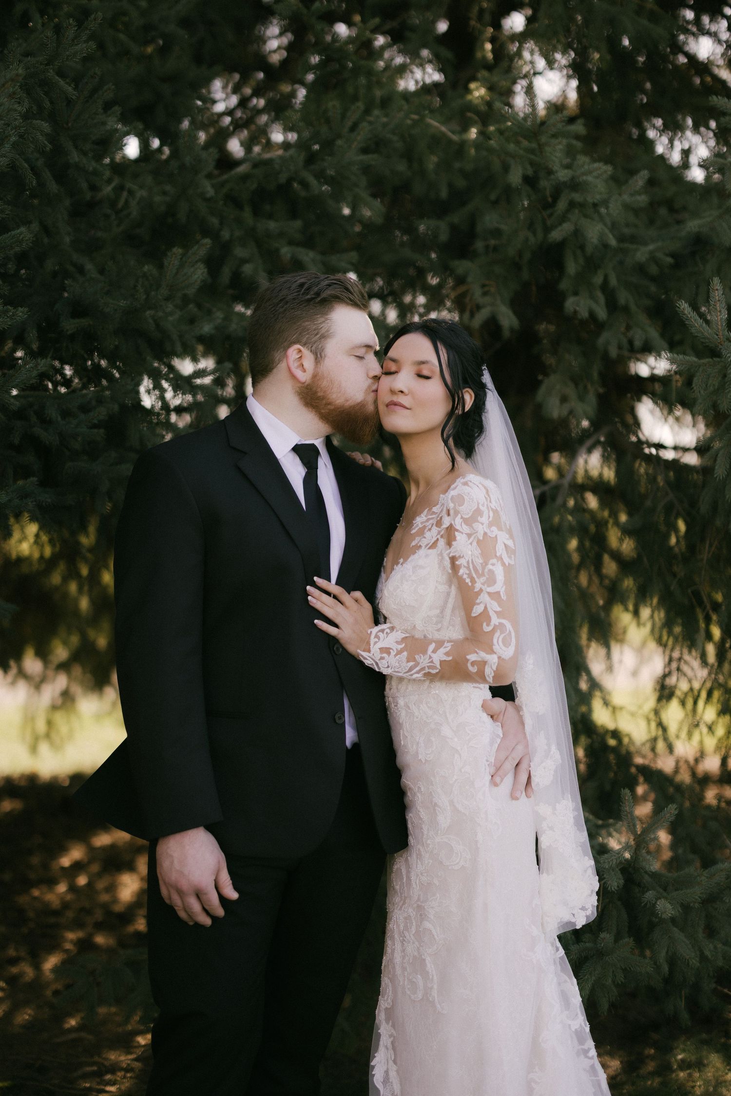 A romantic wedding couple shares intimate moments in a garden setting with evergreen trees in the background.