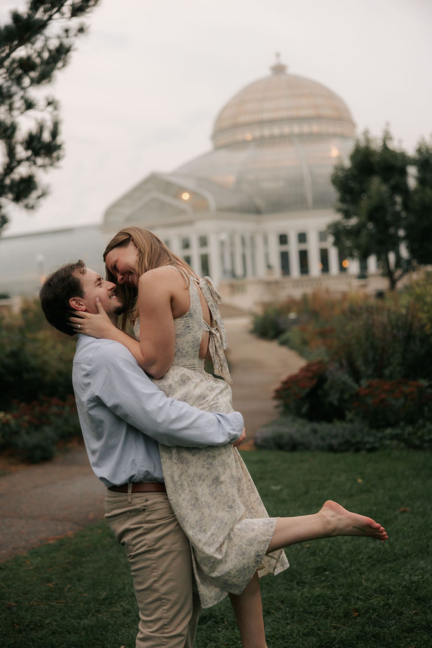 A romantic couple embraces and spins on a grassy lawn with a domed building visible in the background.