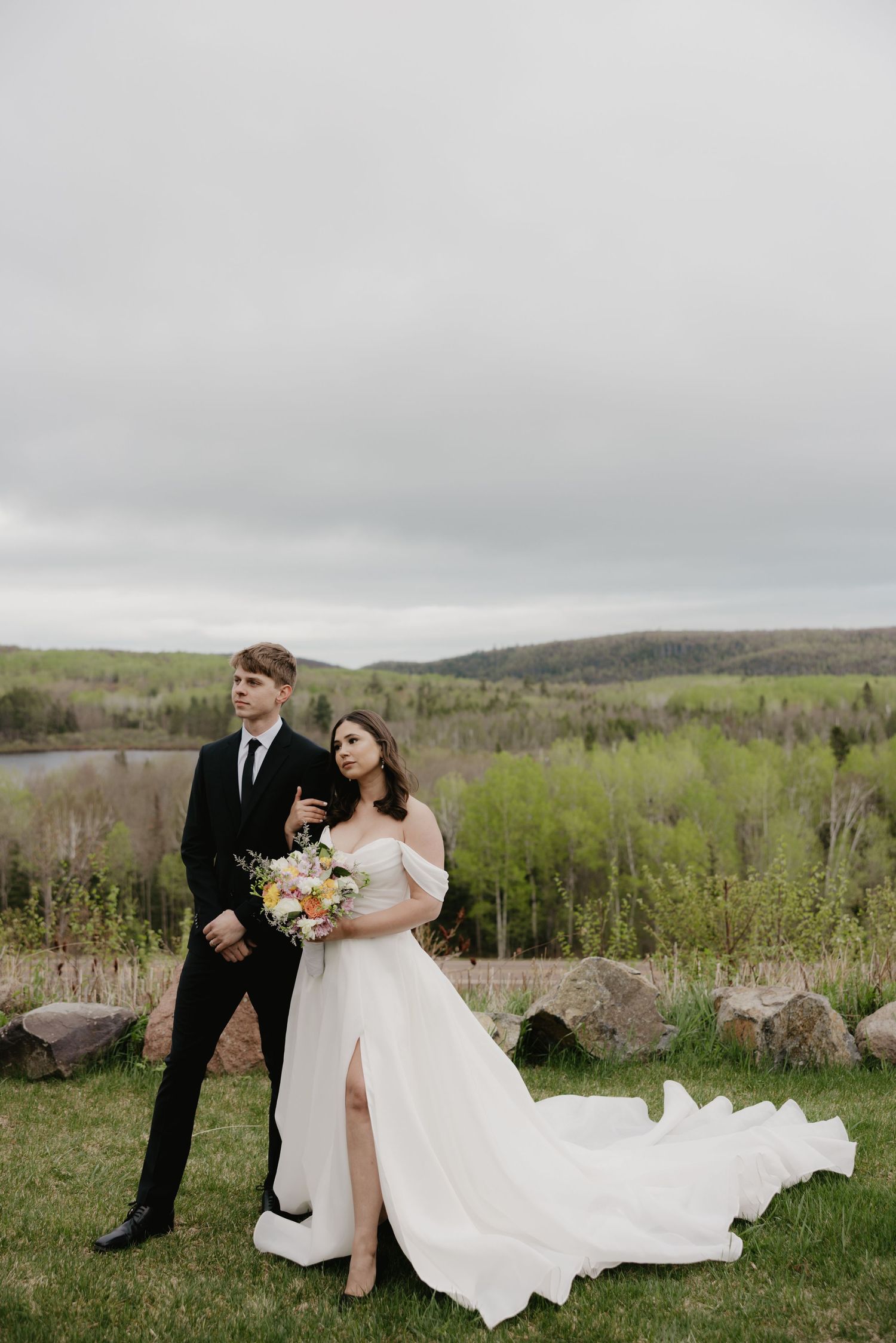 A couple in wedding attire stands on a grassy hilltop overlooking a valley on an overcast day.