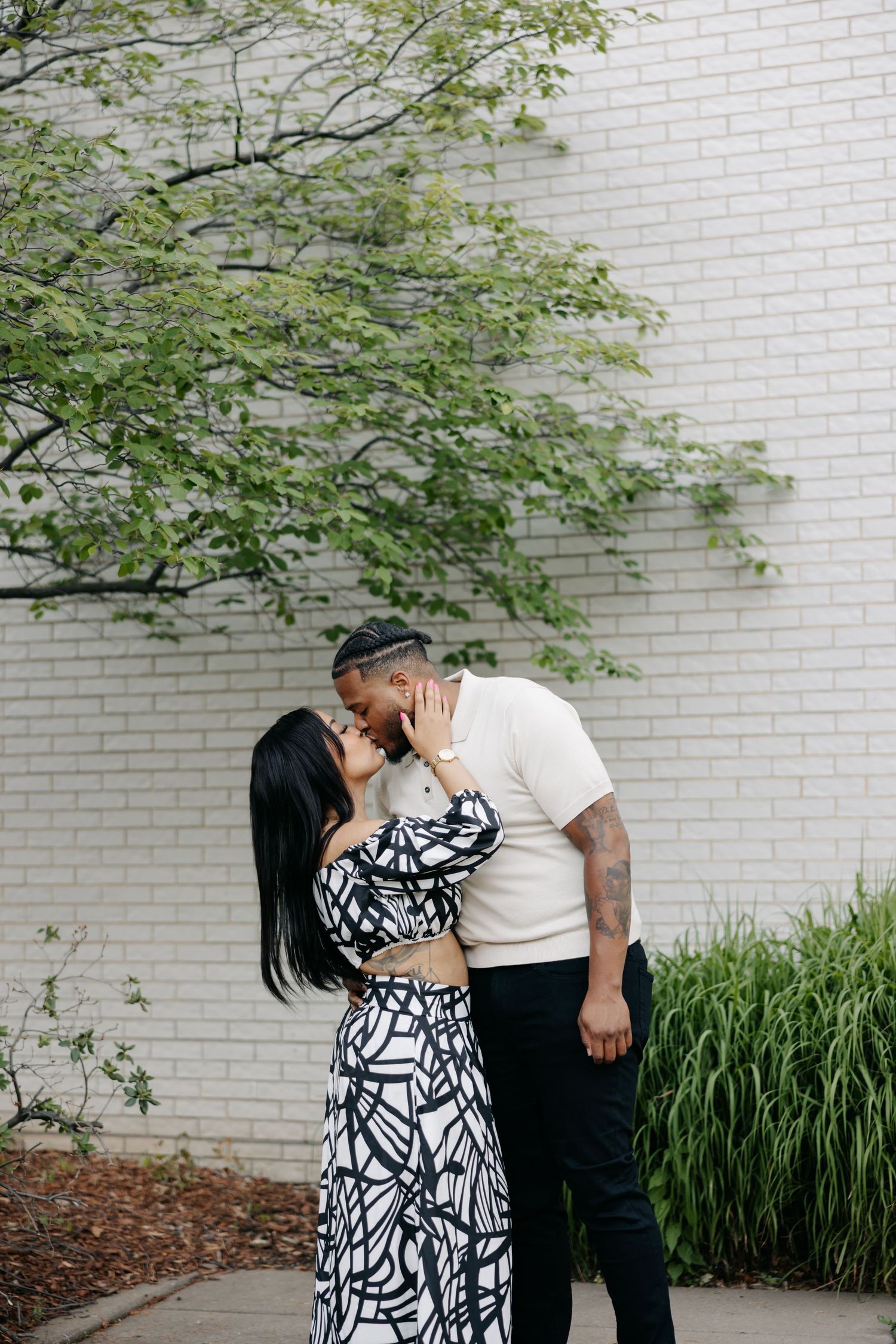 A romantic couple poses against a white brick wall with green foliage, wearing black and white outfits in modern engagement style.