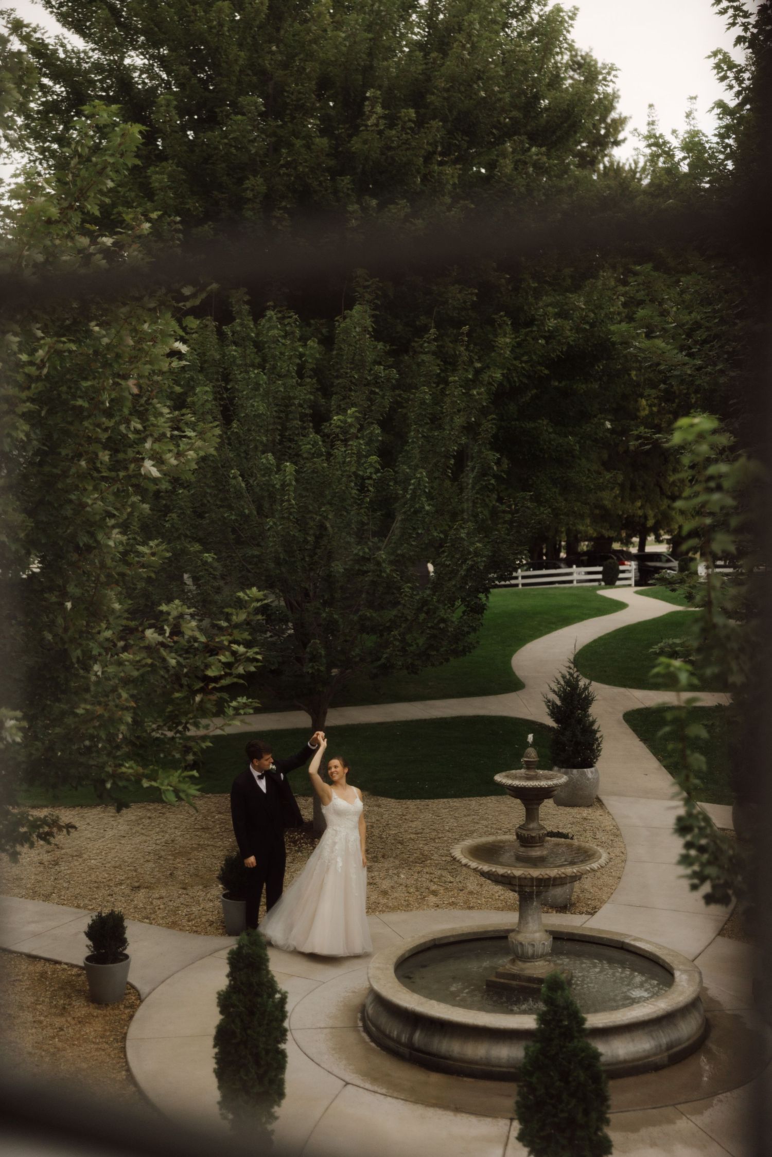 Bride and groom dancing near a stone fountain in a lush garden, captured from above through a window.