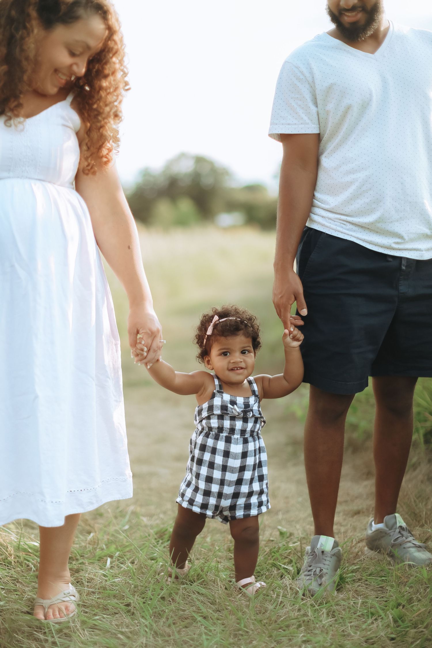 Family holds hands with toddler wearing black and white gingham dress in sunny outdoor field.