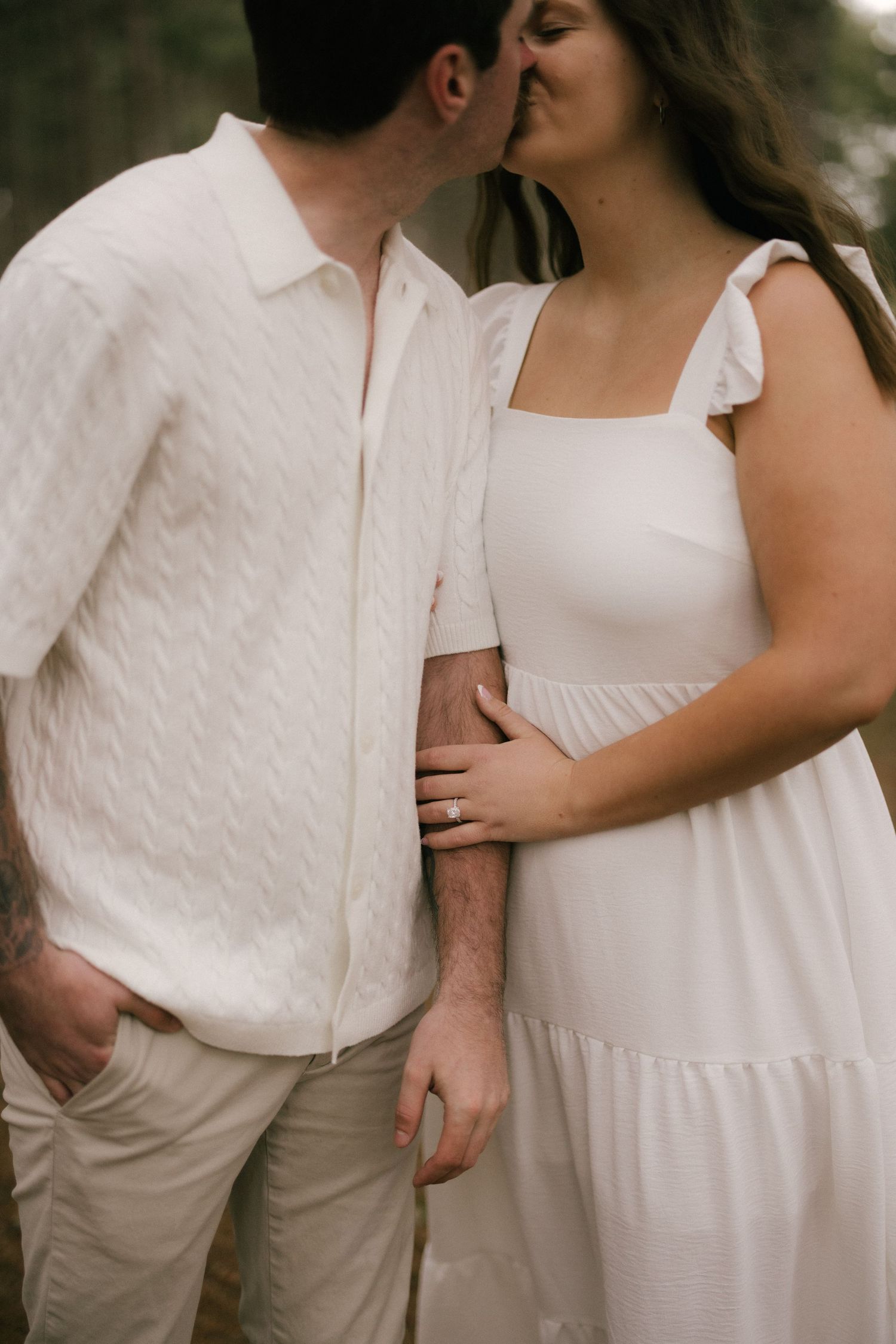 Couple in white outfits sharing a kiss outdoors, woman wearing engagement ring and ruffled sundress.