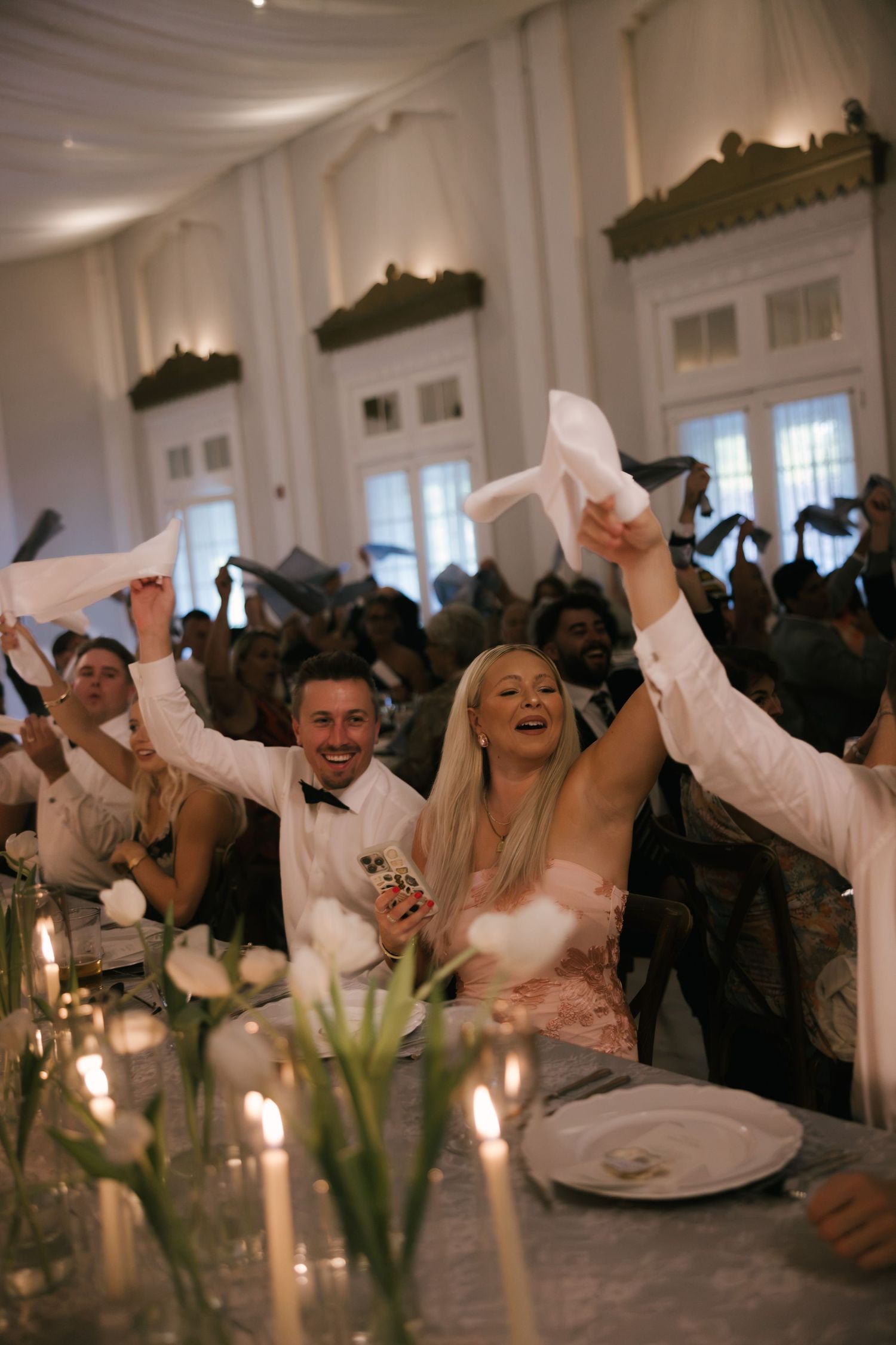 Joyful wedding guests waving white napkins at elegant candlelit reception in grand ballroom.