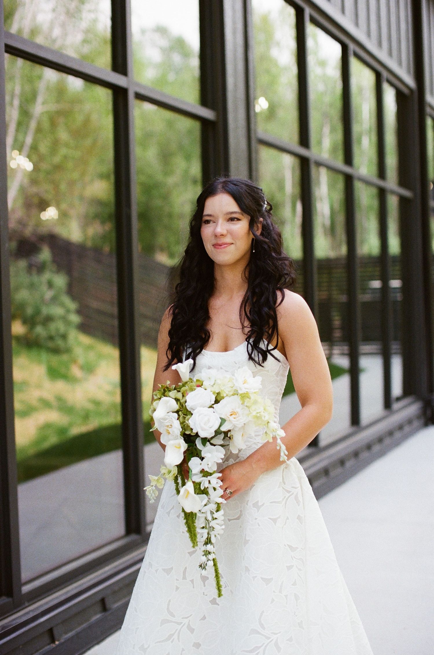 Bride in strapless lace gown holding cascading white orchid bouquet standing before black-framed windows.