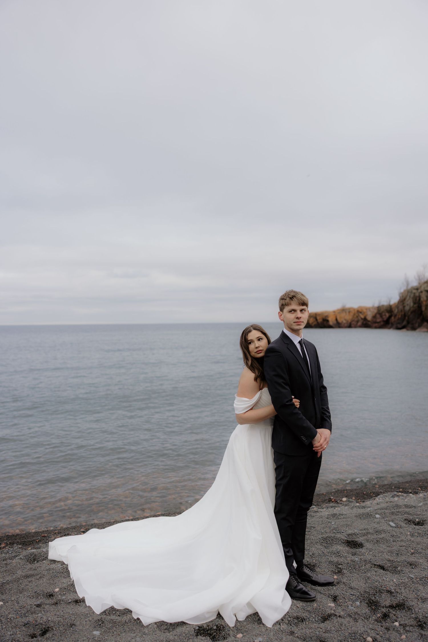 A wedding couple poses on a rocky beach with cloudy skies and water in the background.