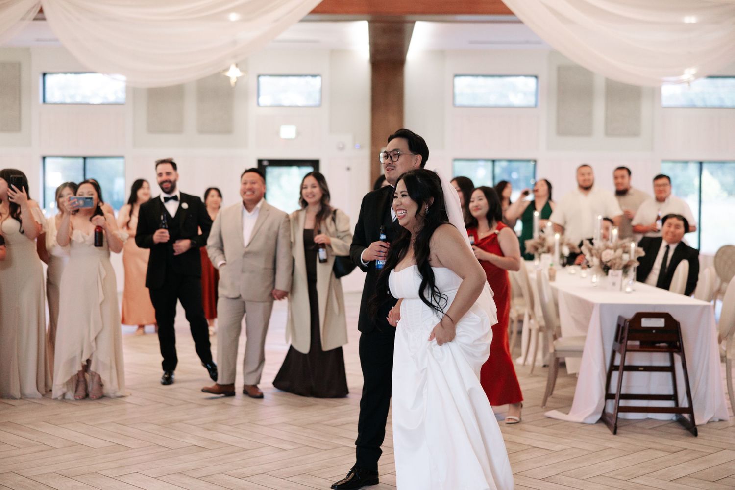 A group of people dancing and celebrating at an elegant white wedding reception venue with large windows.